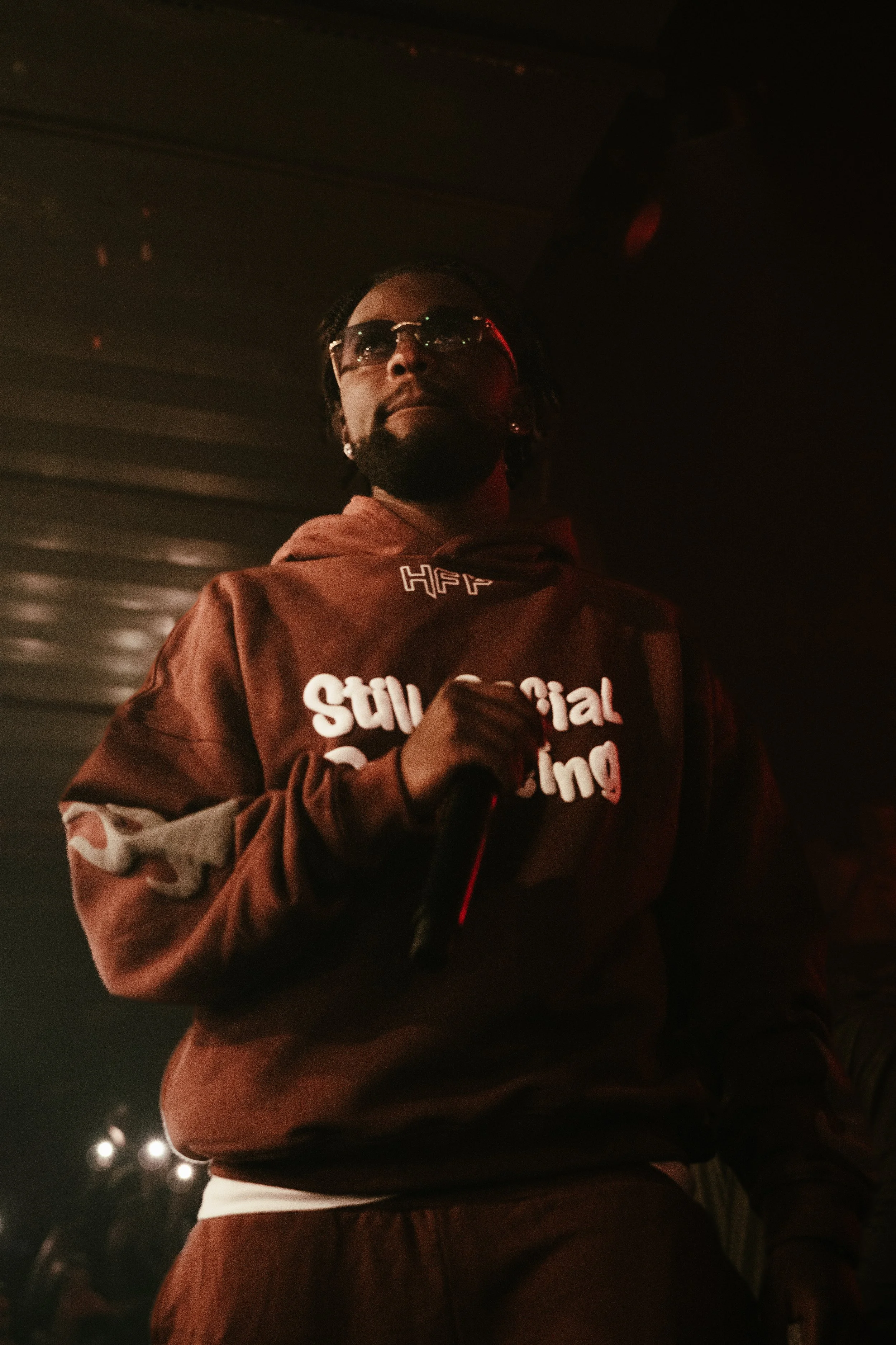 Un homme portant une veste marron avec l'inscription 'Stochastic Rolling' et des lunettes de soleil, tenant un micro dans un environnement sombre, probablement lors d'une performances ou d'un événement.