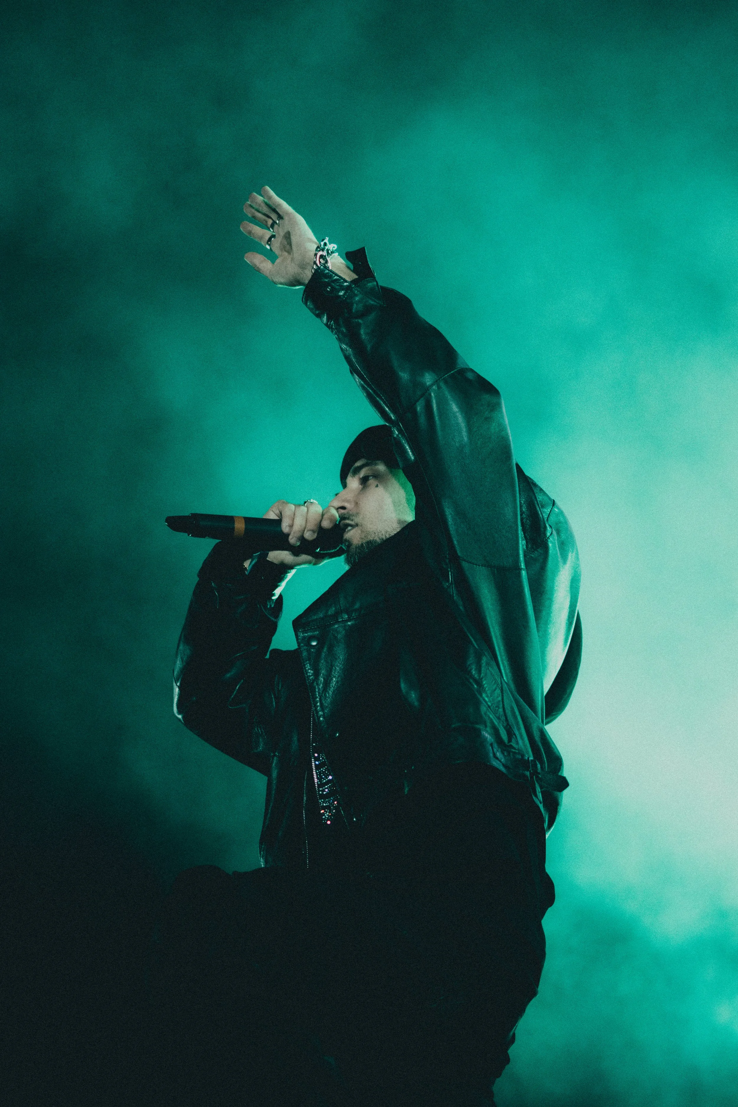 Un homme portant une veste en cuir noire, tenant un microphone et chantant avec un fond de fumée verte.