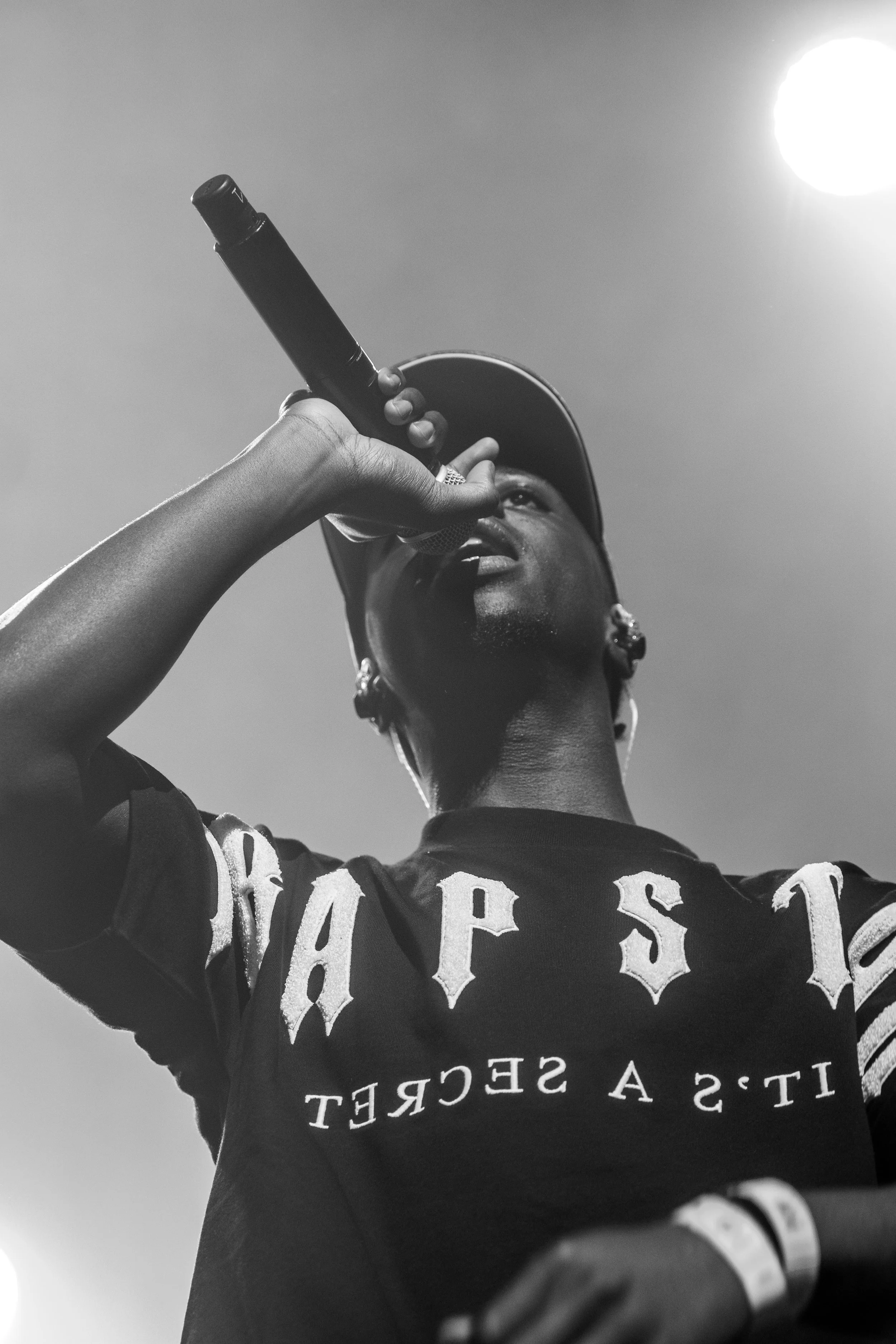 Une personne portant une casquette, tenant un micro, semble chanter ou parler lors d'une performance, vue de dessous avec le soleil en arrière-plan.