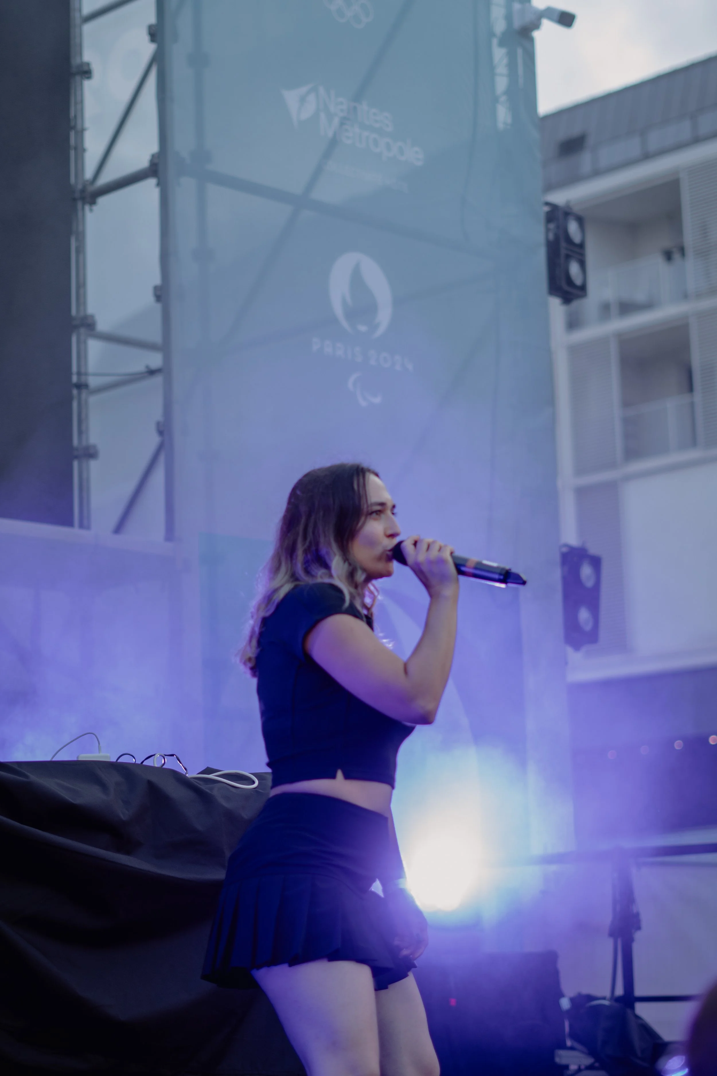 Une jeune femme chante avec un micro lors d'un événement en plein air, avec un fond où l'on peut voir un panneau avec le logo de Nantes Métropole et un symbole indiquant Paris 2024.
