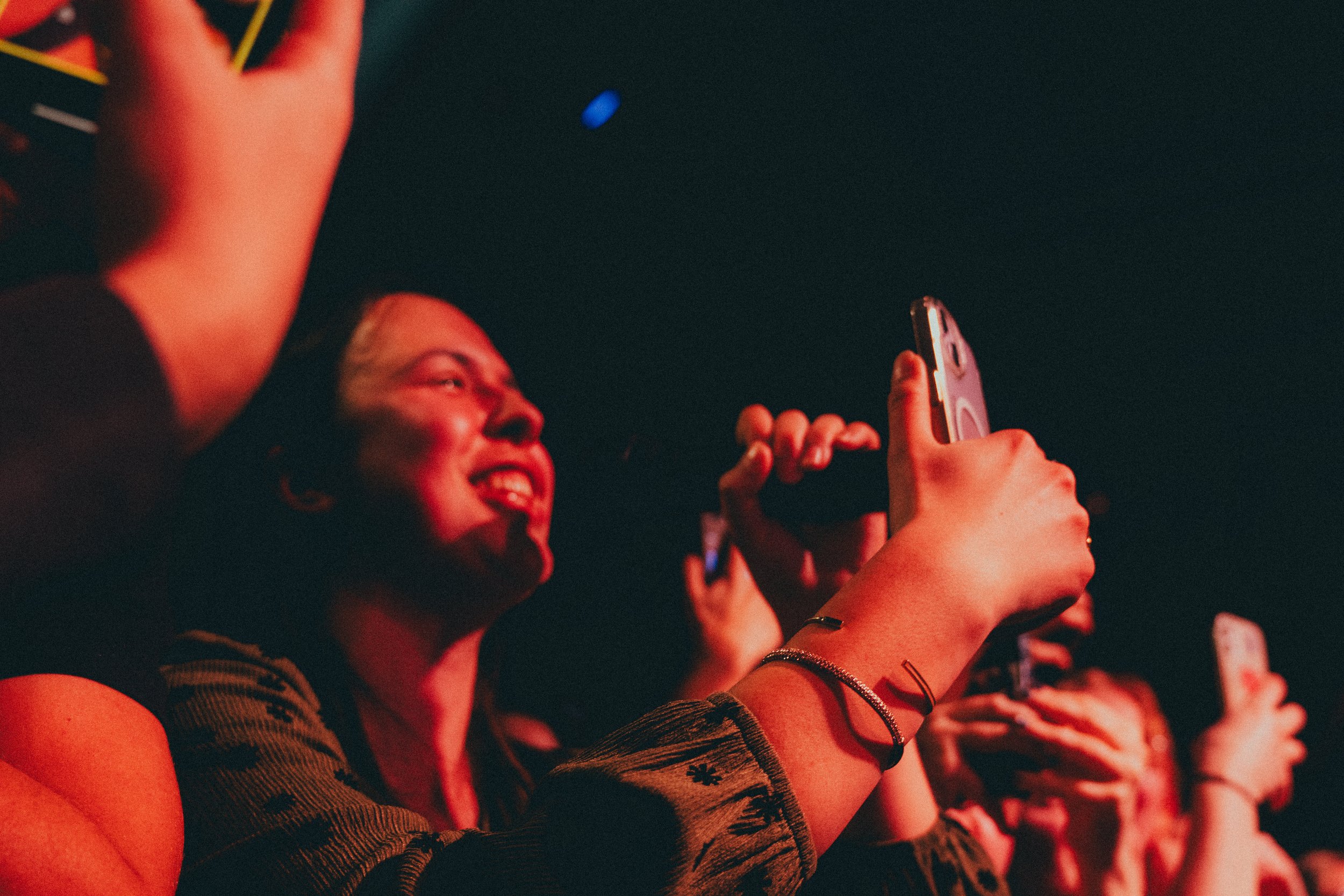 Des jeunes personnes souriantes prennent des selfies ou enregistrement vidéo avec leurs téléphones lors d'un événement ou concert, dans une ambiance sombre et éclairée par des lumières rouges.
