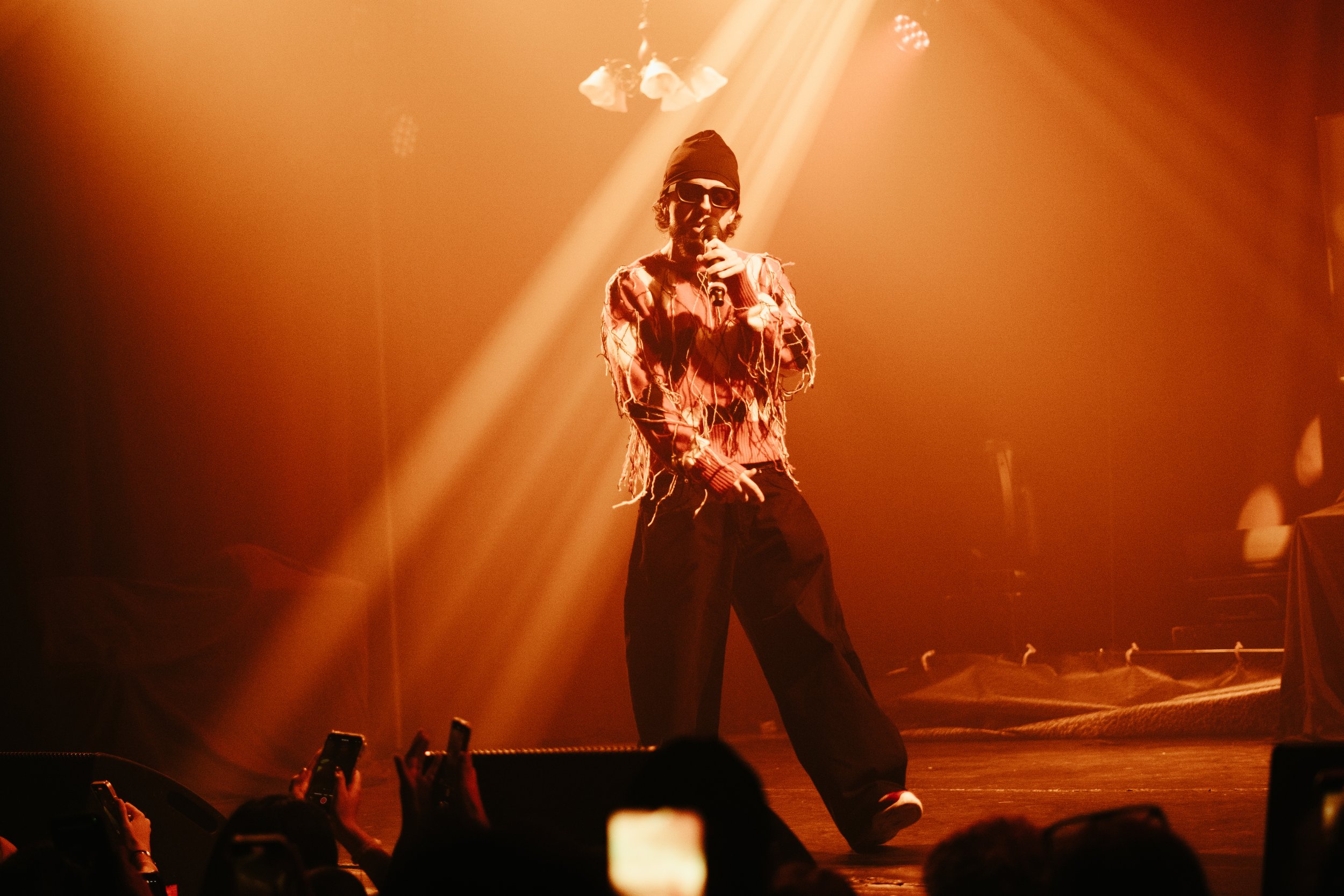 Un artiste en tenue décontractée, portant un bonnet et des lunettes de soleil, chante sur scène sous des lumières chaudes et orangées, avec le public prenant des photos.