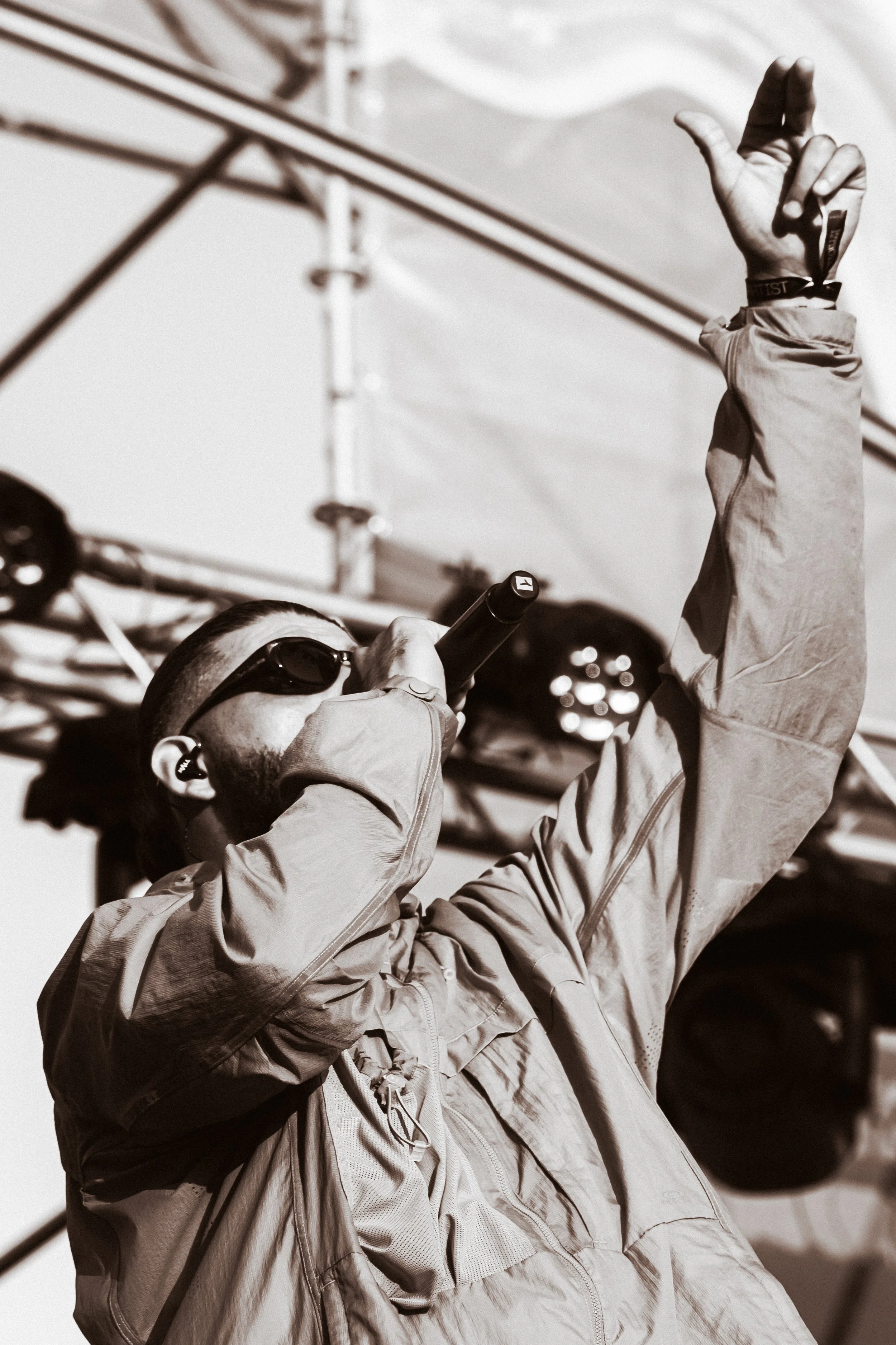 Un homme avec des lunettes de soleil et un casque écoute de la musique en tenant un microphone, avec un bras levé, lors d'une performance en plein air.