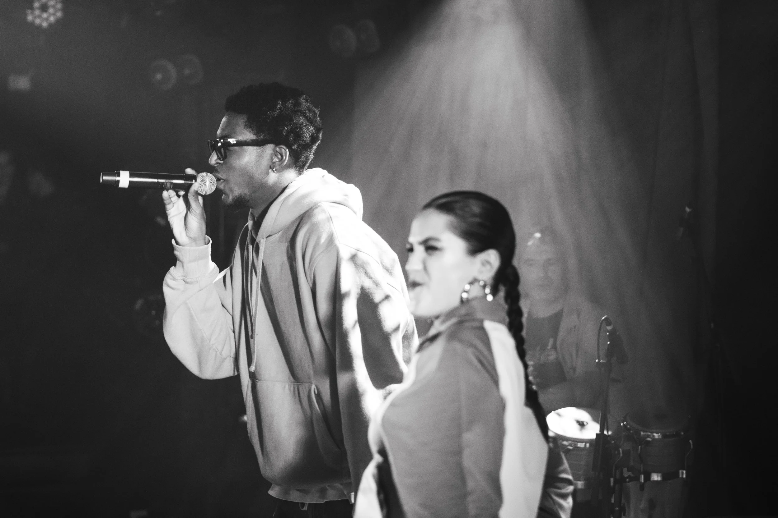 Un homme noirs avec des lunettes et un hoodie blanc chante dans un microphone, une femme à côté avec les cheveux en braids, dans un environnement de scène avec des lumières et des musiciens en arrière-plan.