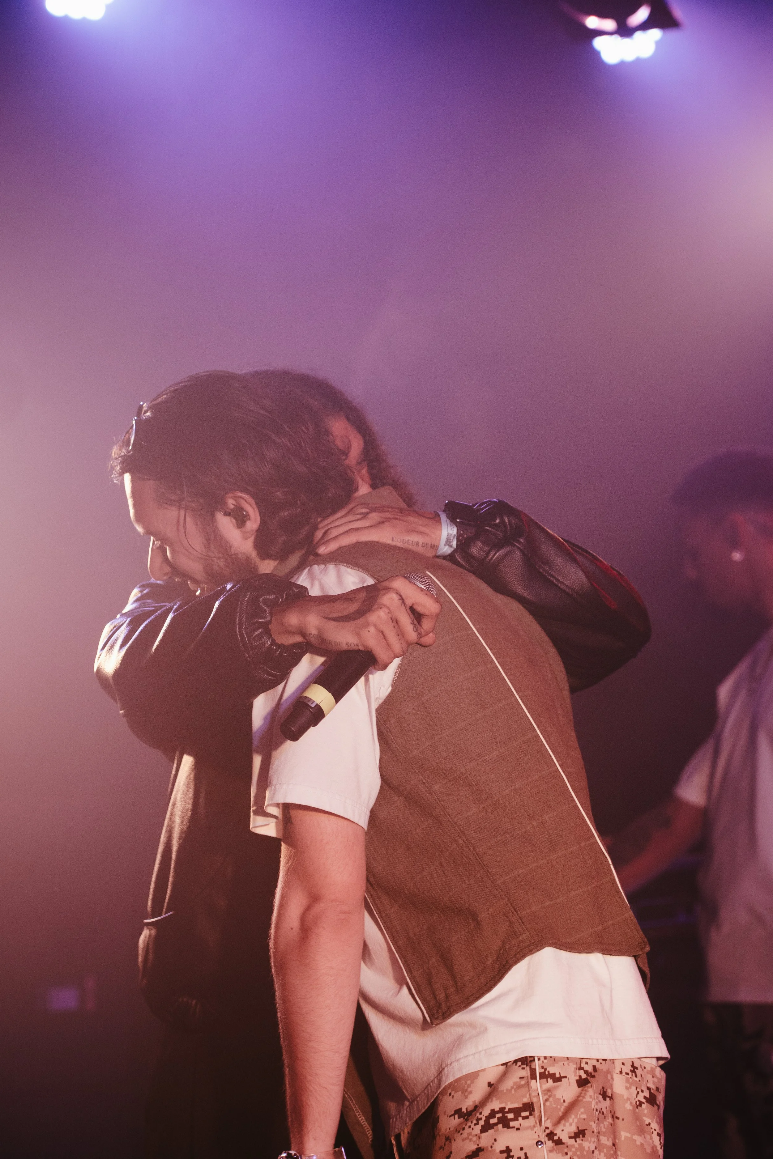 Deux personnes s'embrassent sur scène lors d’un concert, une tient un microphone et la scène est éclairée par des lumières violettes.
