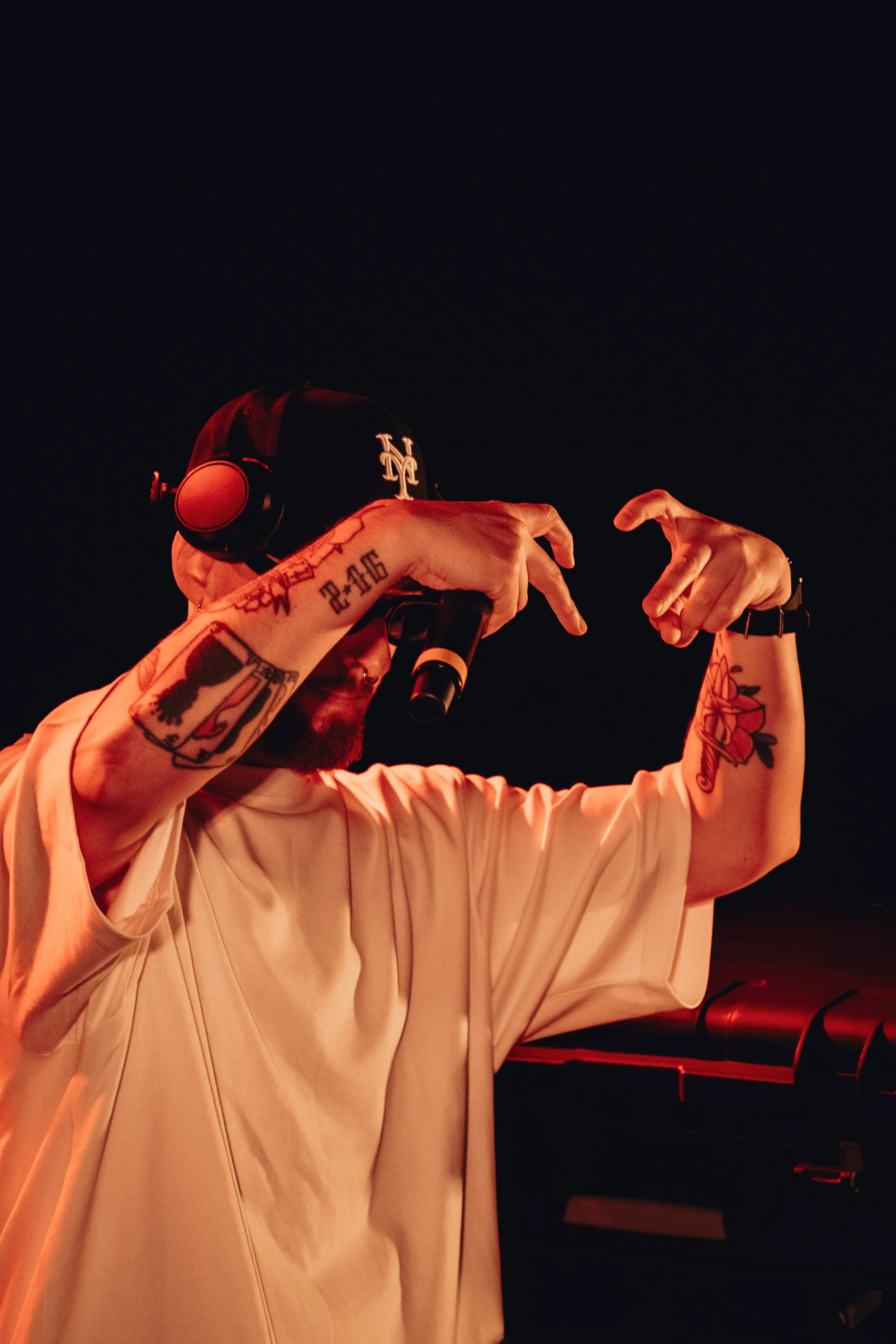 Un homme avec des tatouages, portant un casque avec un logo, joue de la musique ou chante devant un microphone dans un environnement sombre et chaud.