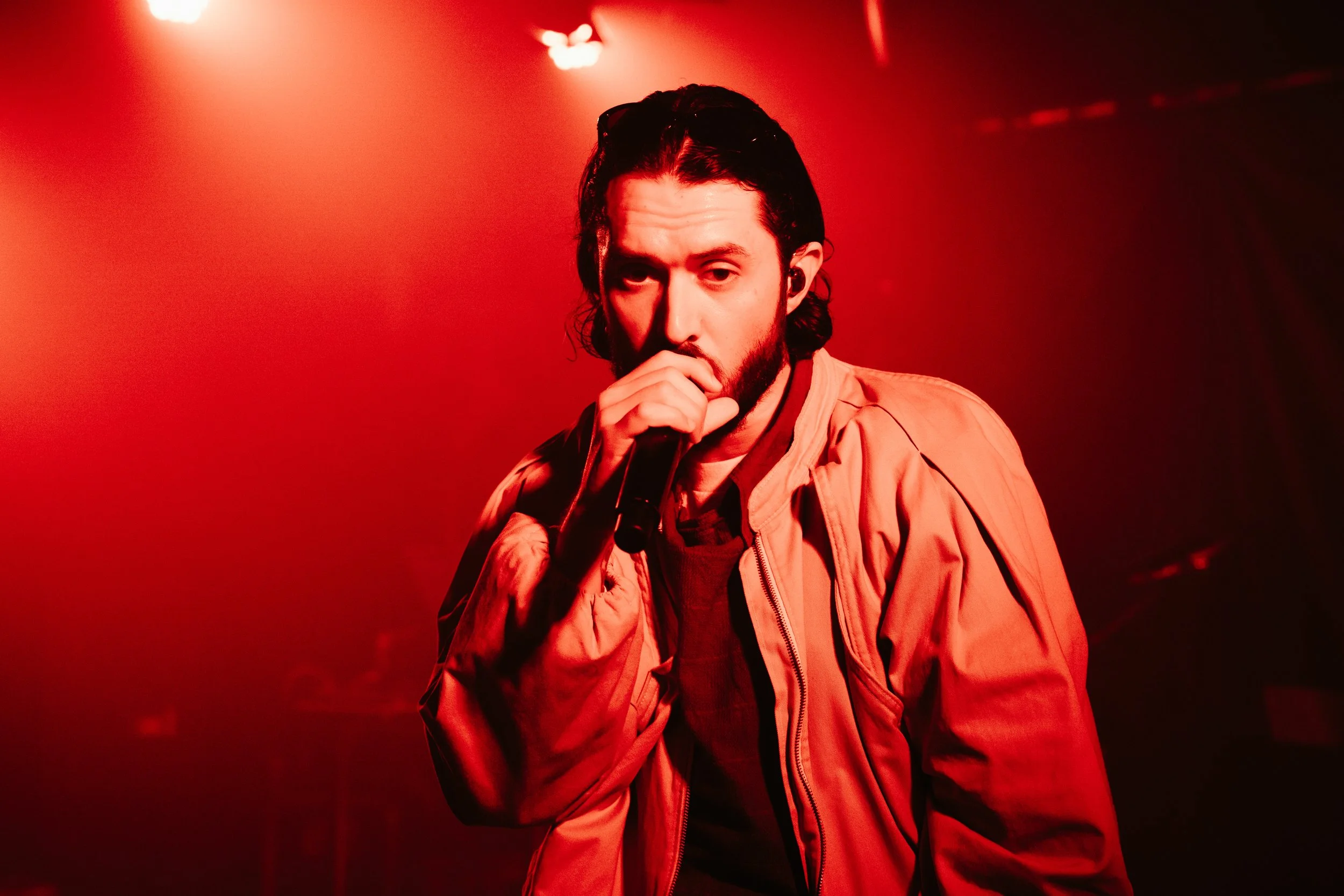 Un homme avec des cheveux longs et une barbe, tenant un microphone, dans un éclairage rouge, probablement lors d'une performance musicale.