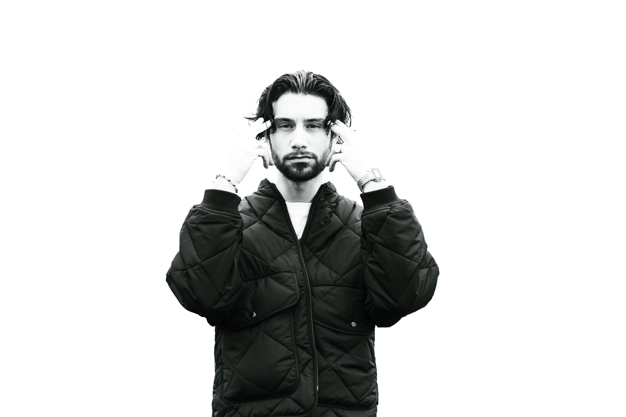 Jeune homme avec barbe et cheveux longs, portant un blouson matelassé, tient ses mains près de sa tête, regard sérieux, fond blanc