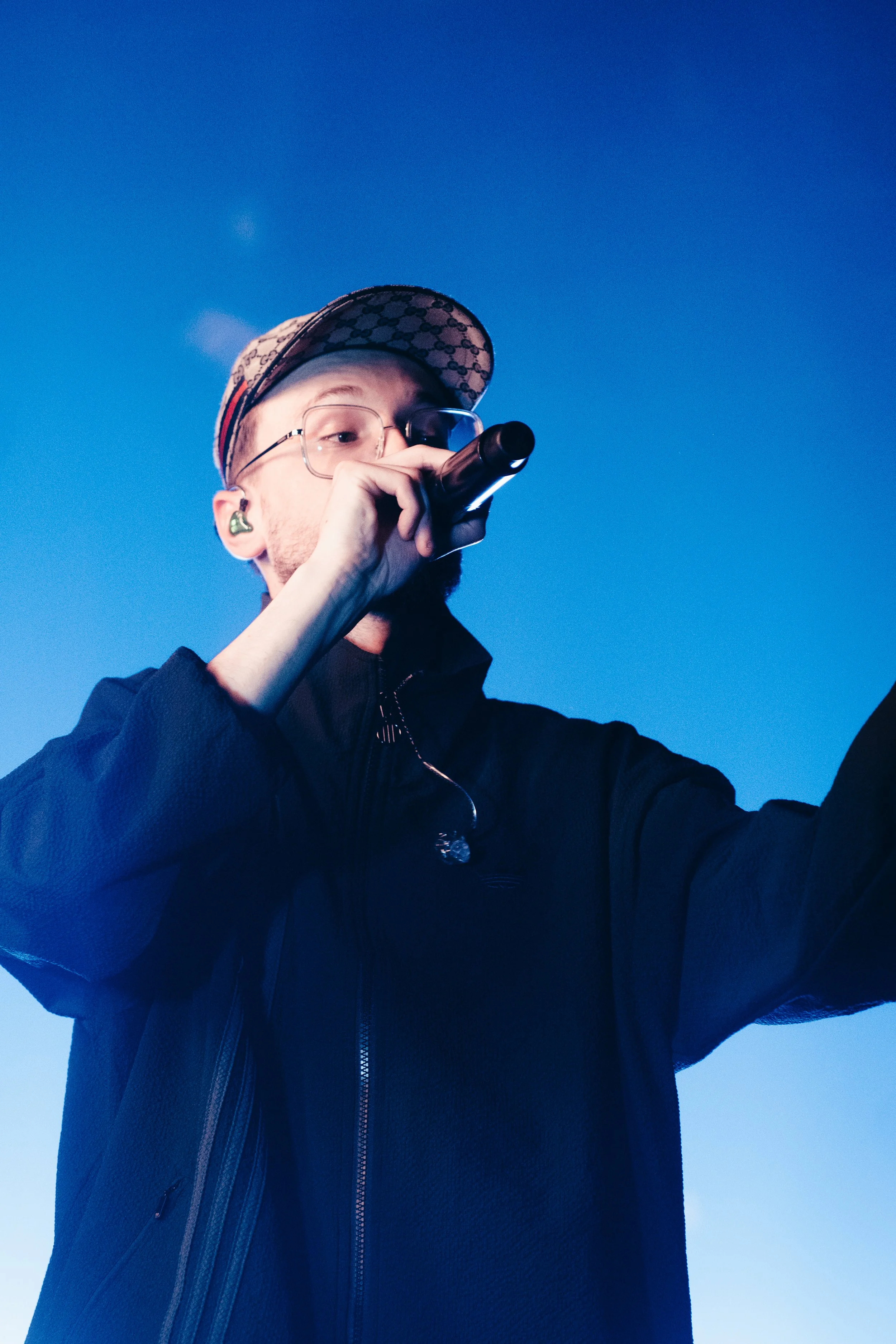 Un jeune homme portant une casquette, des lunettes et un casque d'écoute, tient un microphone et fait un selfie contre un ciel bleu.