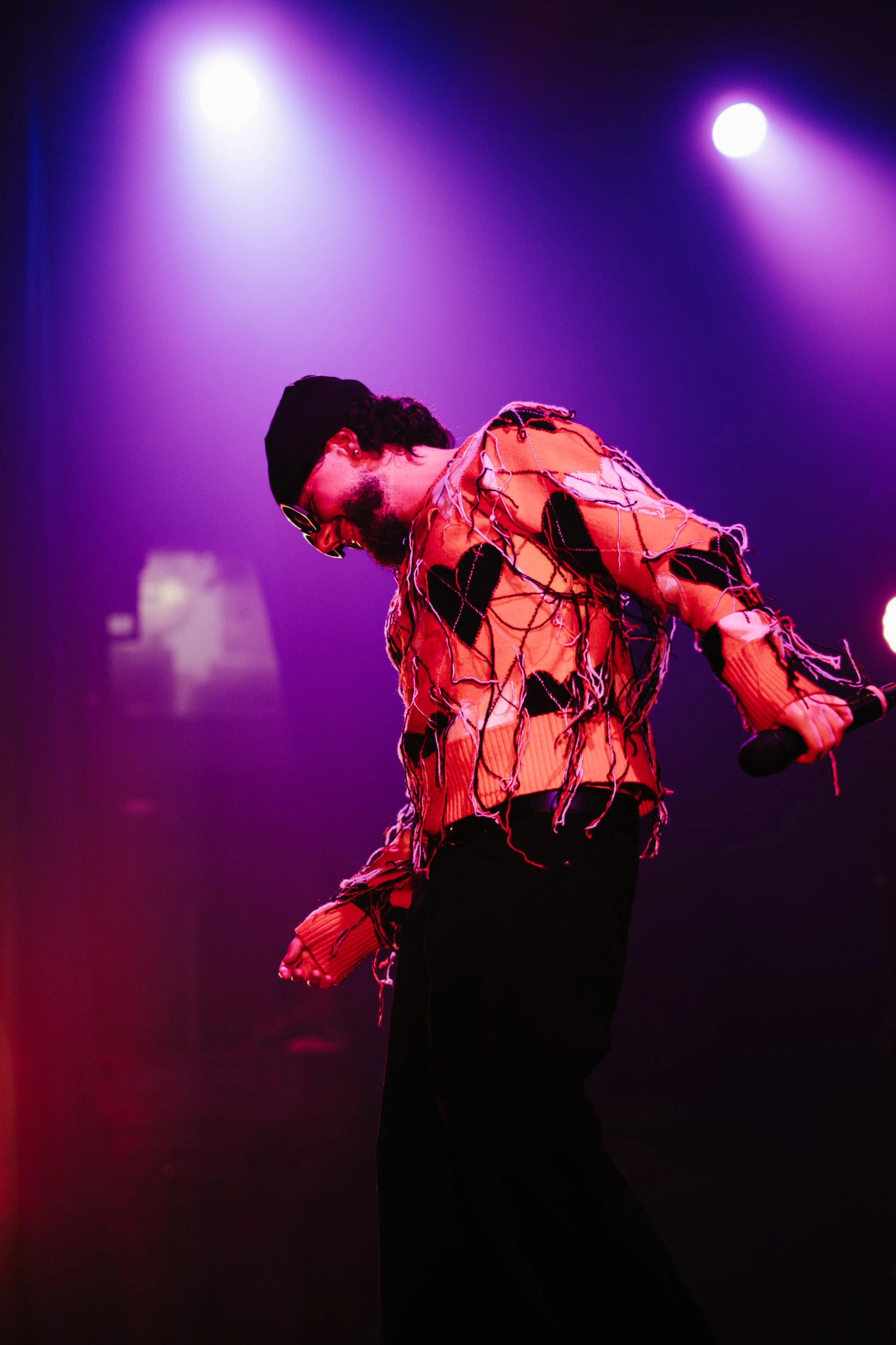 Un homme portant un sweat-shirt orange déchiré avec des fils suspendus, casquette noire, lunettes de soleil, tenant un micro, sous des projecteurs violets et bleus.