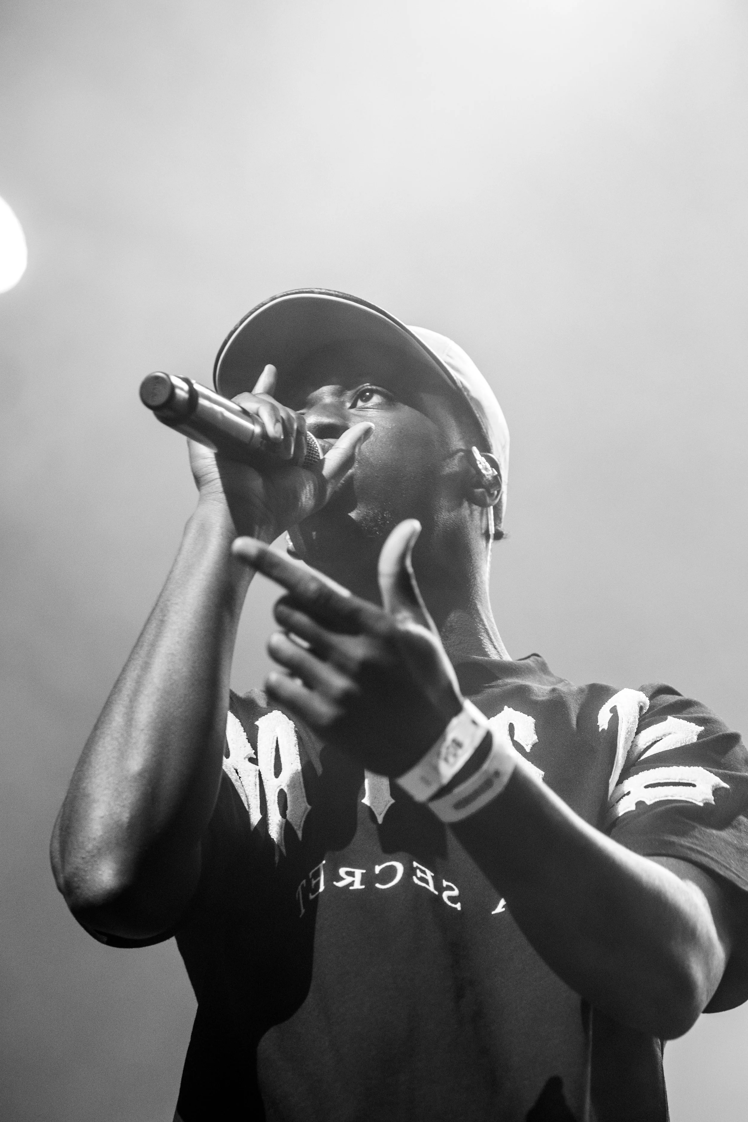 Un homme noir portant une casquette et un t-shirt noir chante ou parle dans un micro, en noir et blanc.