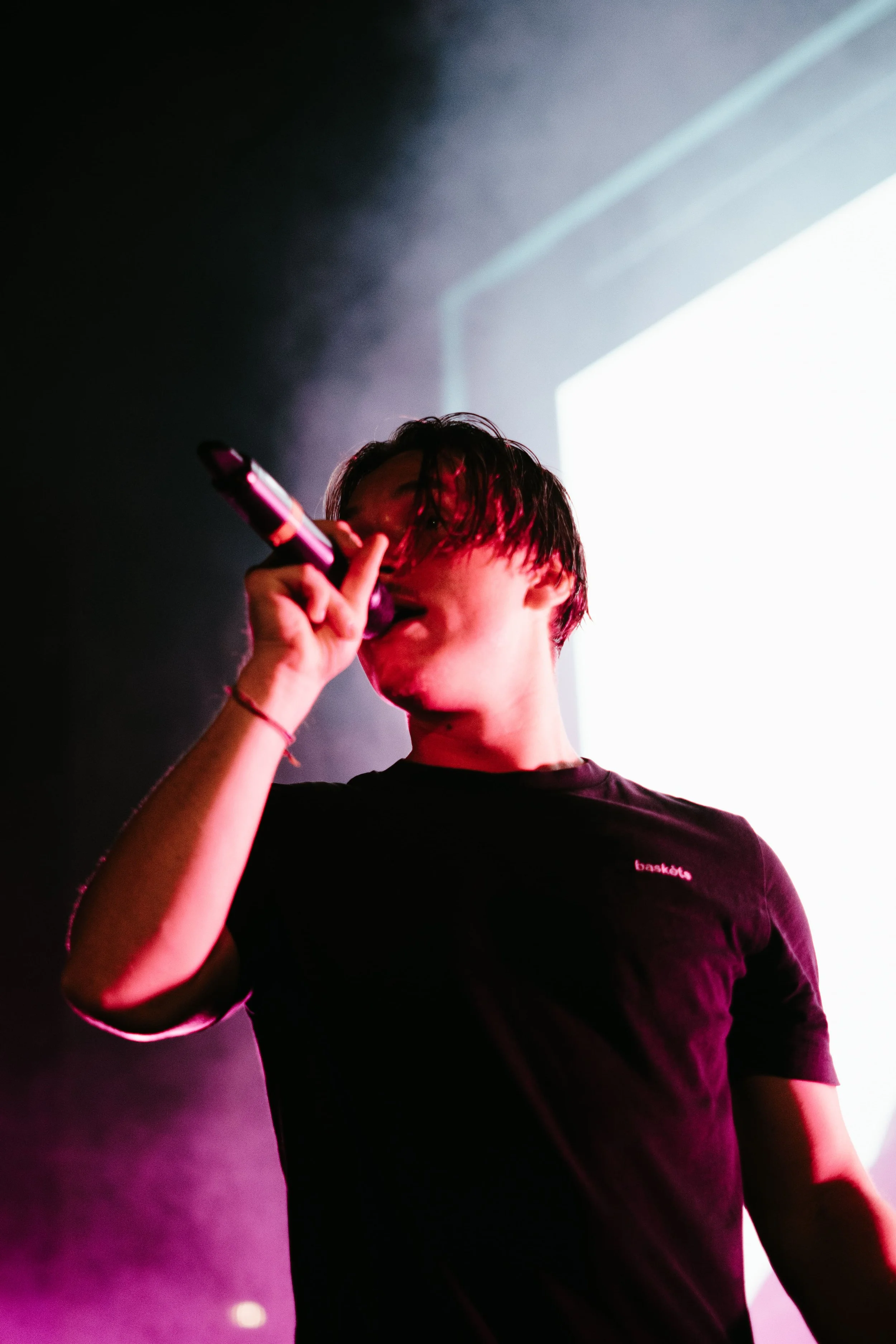 Jeune homme chantant ou parlant dans un microphone lors d'une performance sur scène, avec un fond éclairé en blanc et une lumière rose ou violette.