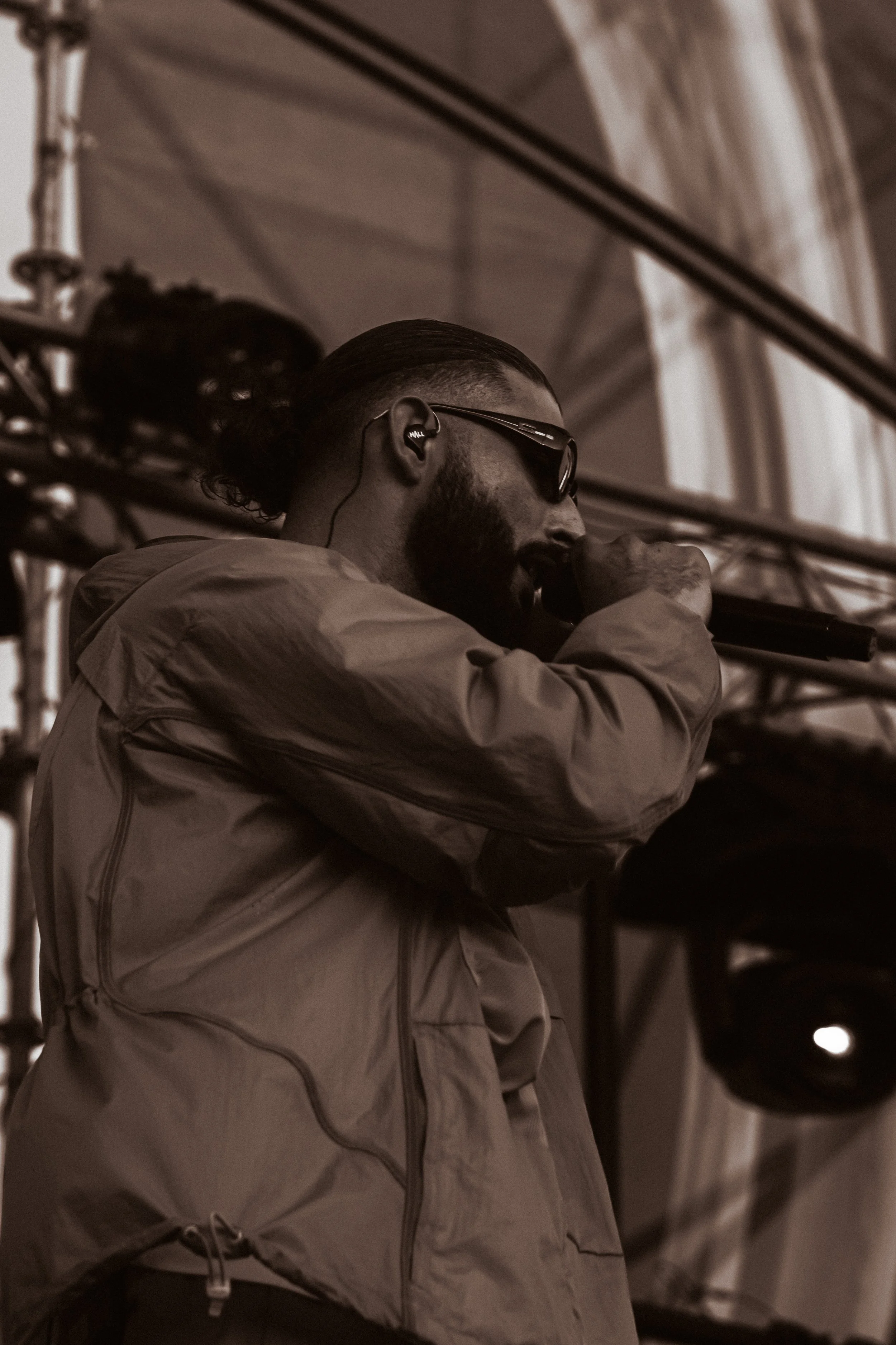 Un homme portant des lunettes de soleil et un casque d'écoute, tenant un microphone lors d'une performance ou d'un concert, avec une structure métallique visible en arrière-plan.
