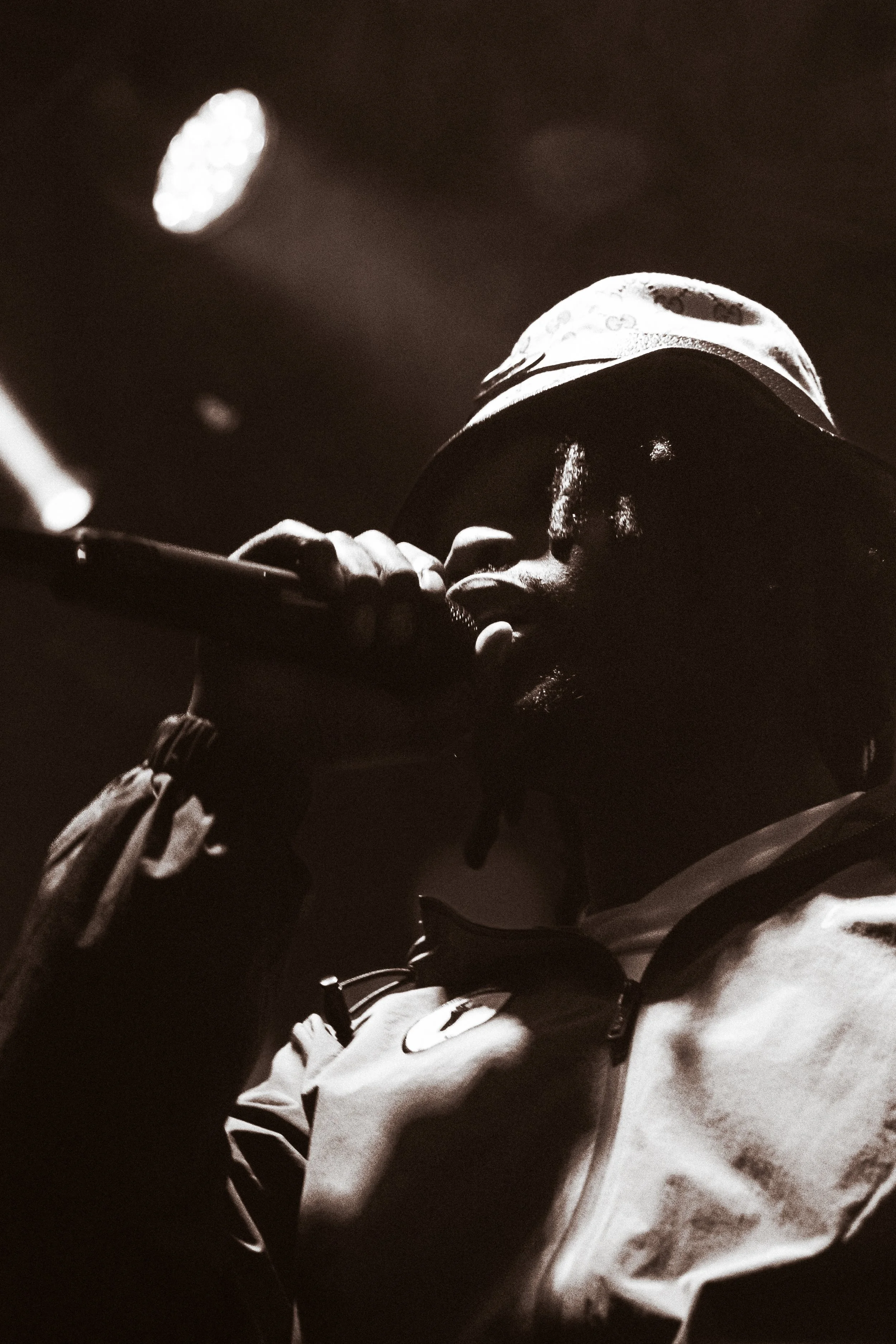 Une personne portant une casquette et un blouson, tenant un micro près de la bouche, en concert ou en performance, en noir et blanc.