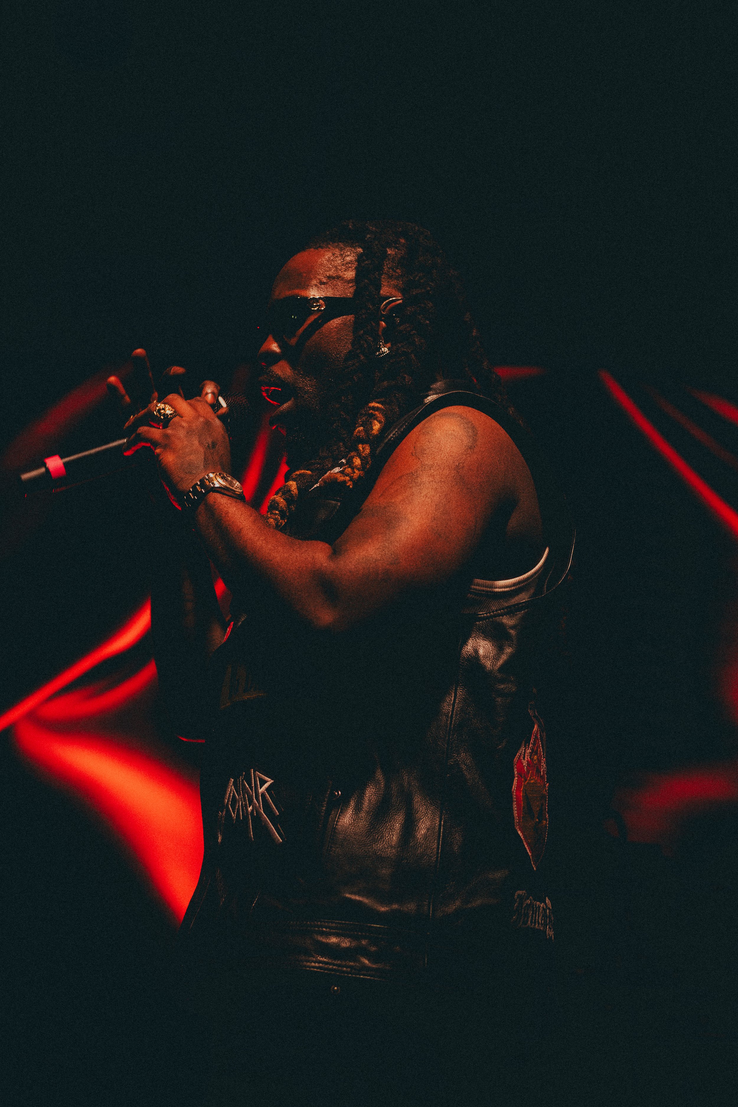 Un homme avec des cheveux longs et tressés, portant des lunettes noires, tenant un micro. Il porte un gilet en cuir avec des patches, dans une atmosphère sombre avec des lumières rouges en arrière-plan.
