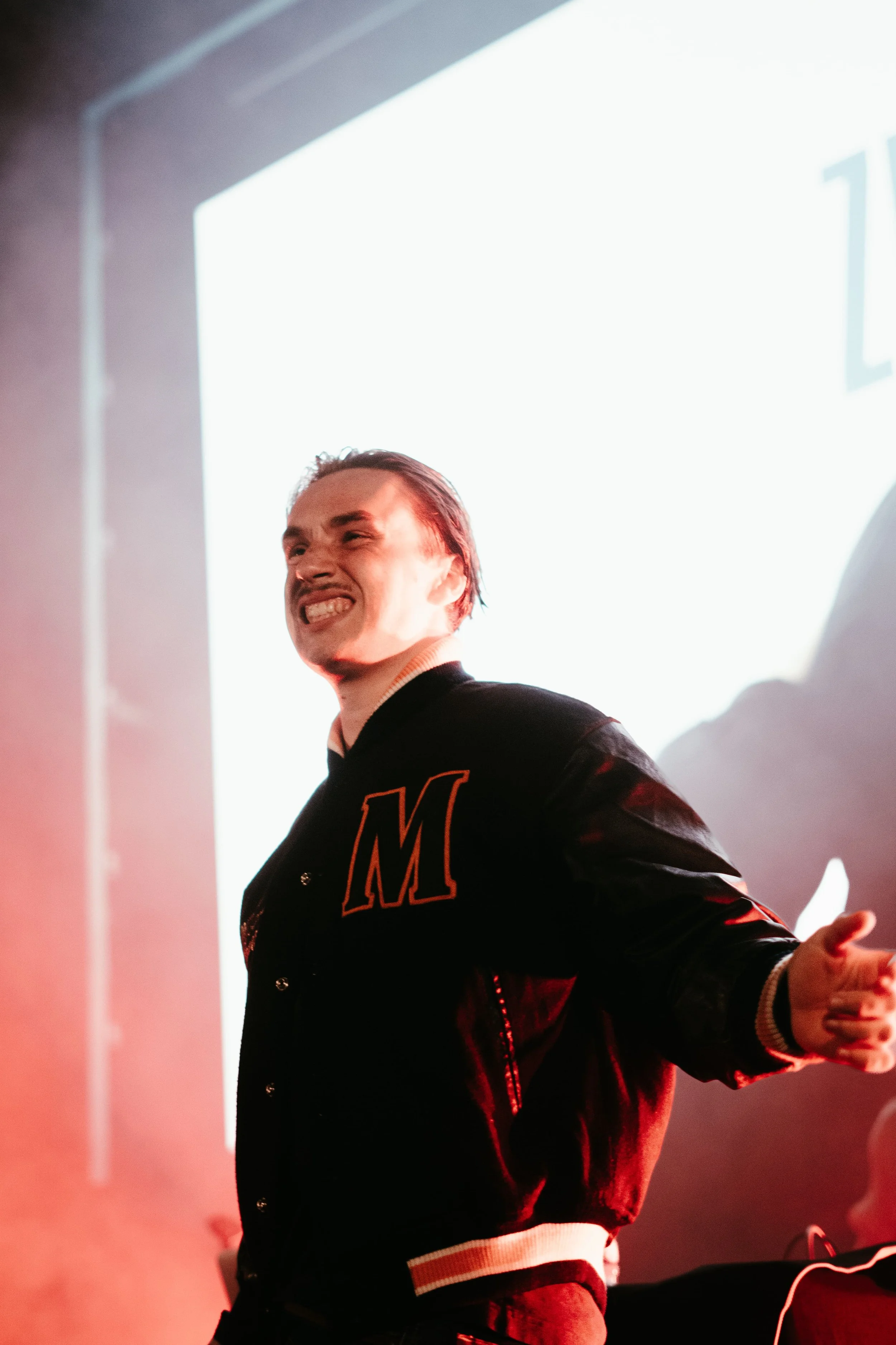 Un homme souriant portant un blouson avec la lettre 'M' sur la poitrine dans une salle avec un écran lumineux en arrière-plan.