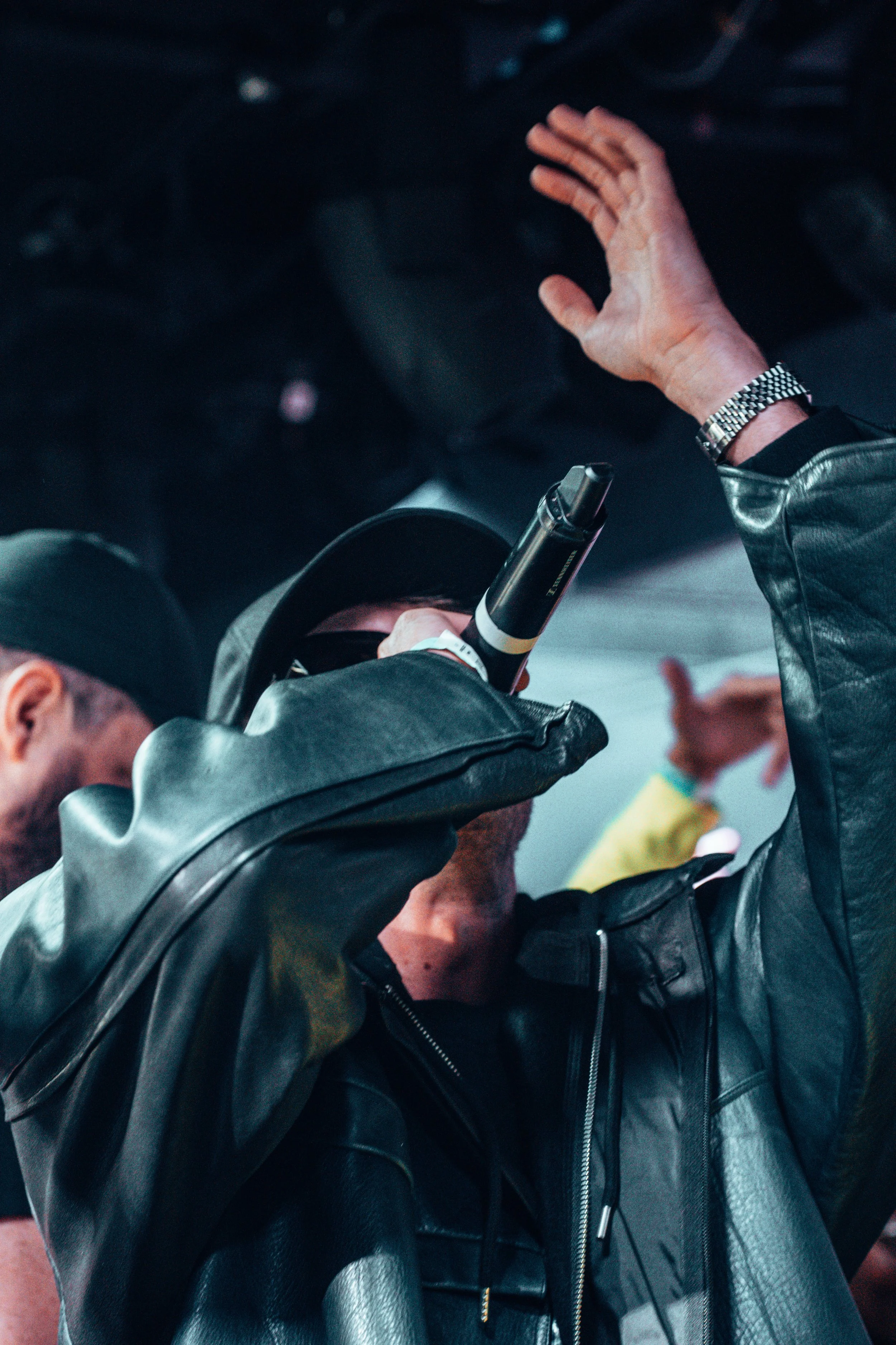 Un homme portant un chapeau noir, des lunettes de soleil, une veste en cuir noire, et un bracelet en métal, chante ou parle dans un microphone lors d'une performance ou d'un événement.