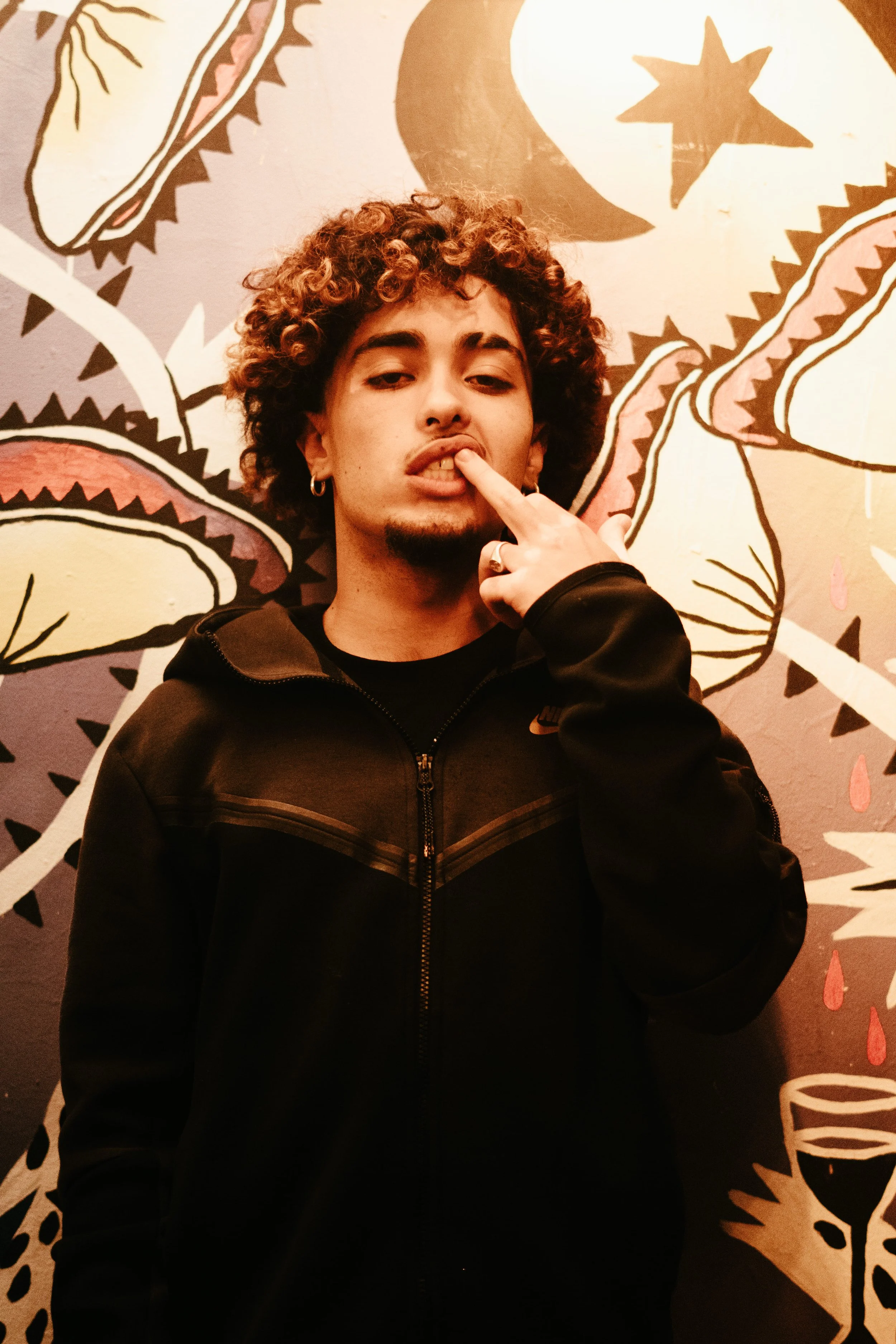 Jeune homme avec cheveux frisés, portant un sweat noir, faisant un geste de finger pistols près de la bouche, posé devant un mur de décoration artistique avec des formes abstraites et des étoiles.