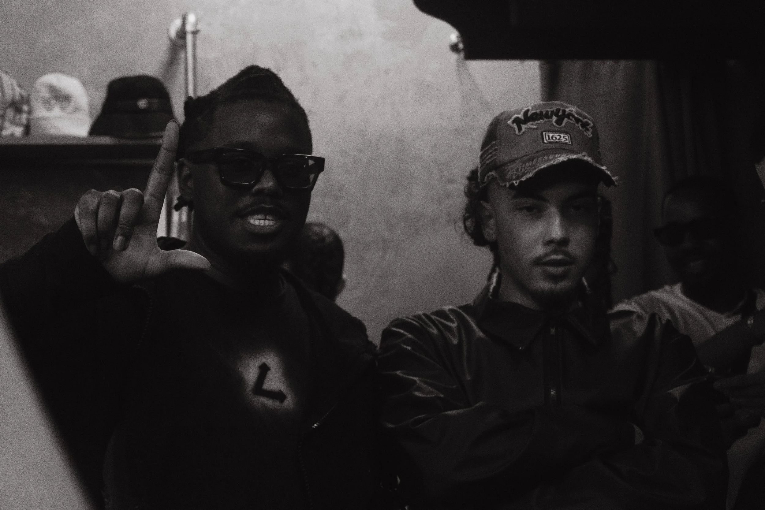 Trois jeunes hommes en intérieur, deux d'entre eux regardent l'objectif. L'un porte des lunettes de soleil et montre un signe de main, un autre porte une casquette et regarde droit devant. L’éclairage est faible, et la photo est en noir et blanc.