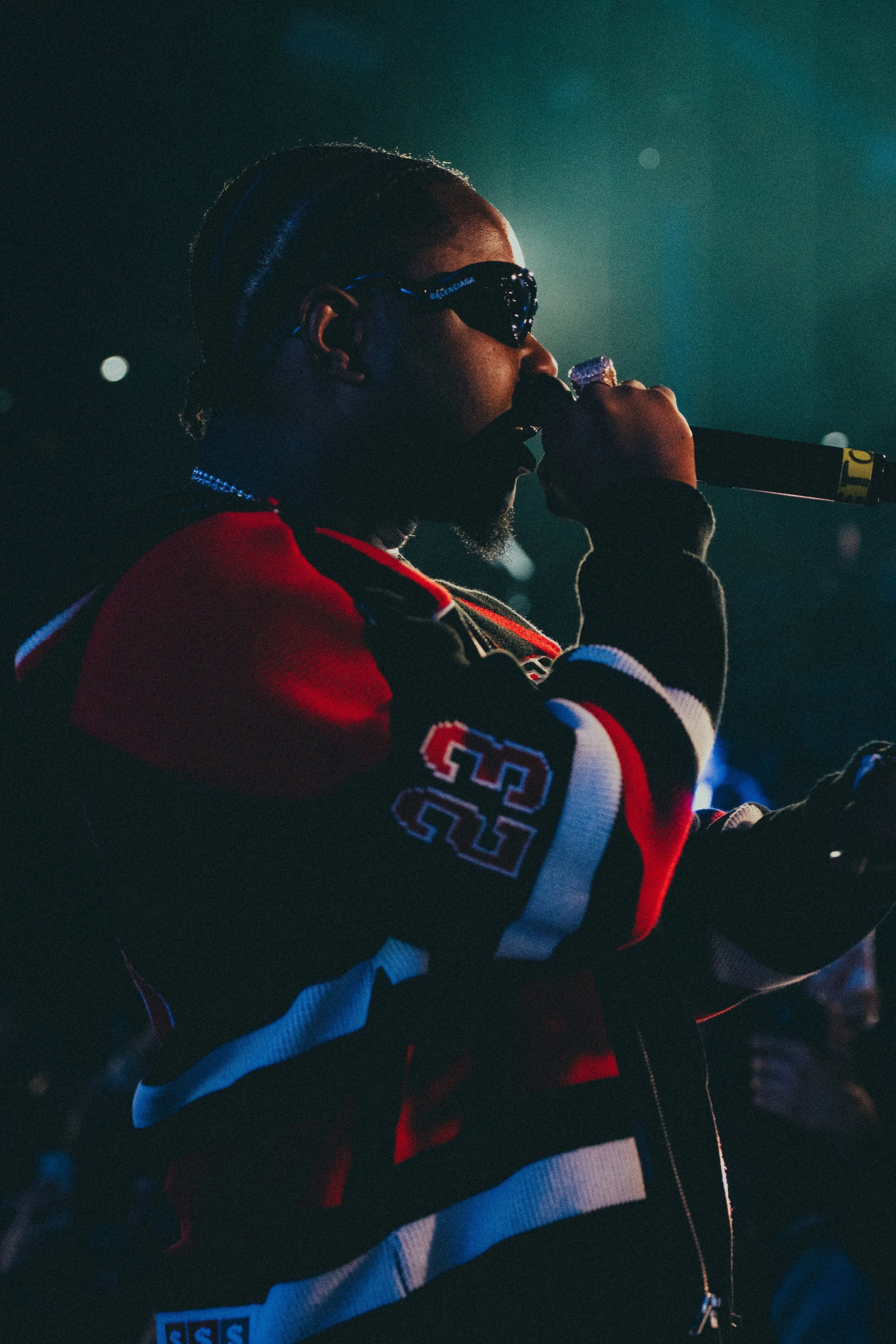 Une personne portant des lunettes de soleil et un sweat-shirt coloré, tenant un micro, en performance sur scène.