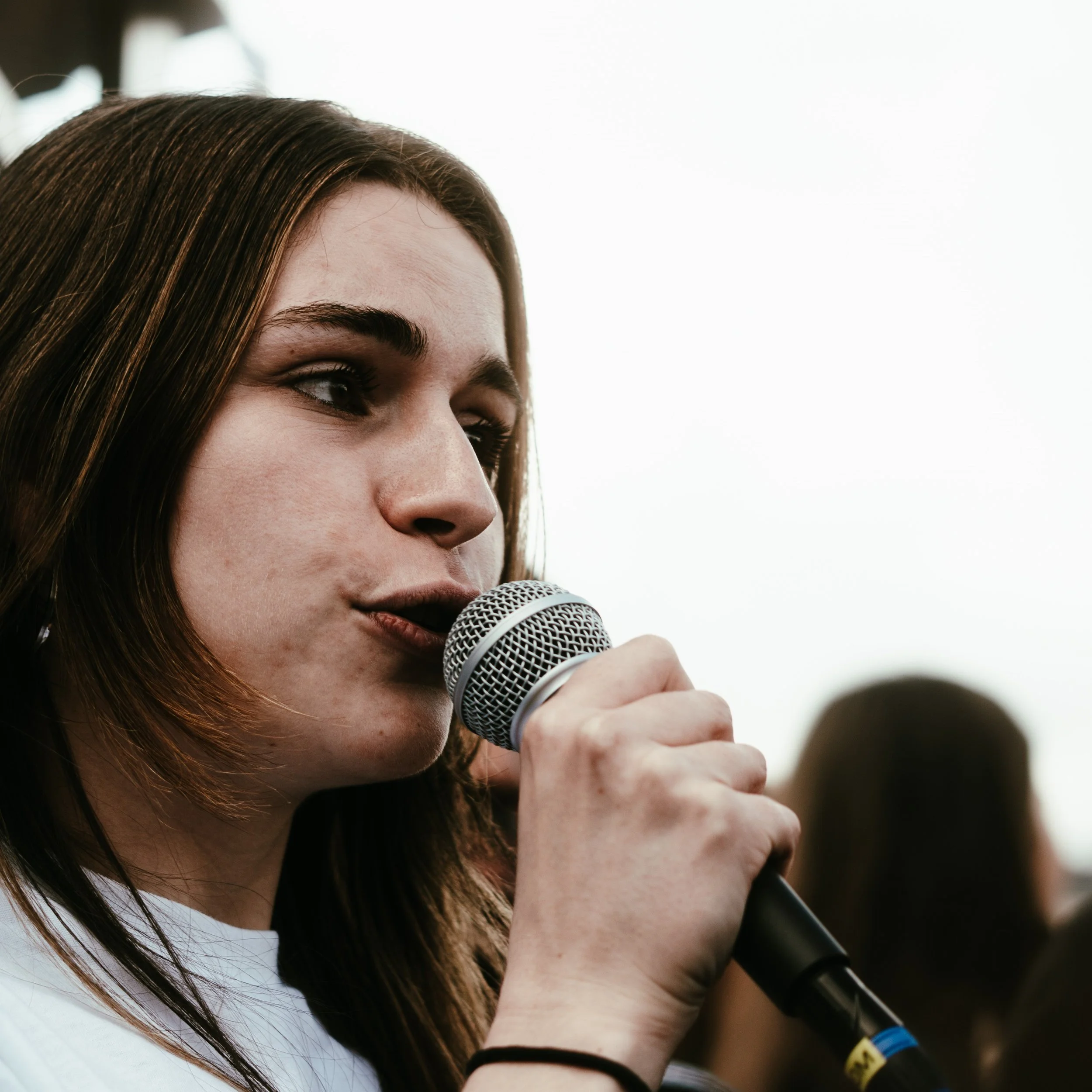 Une jeune femme avec les cheveux bruns, utilisant un microphone lors d'un rassemblement ou d'un événement en plein air.