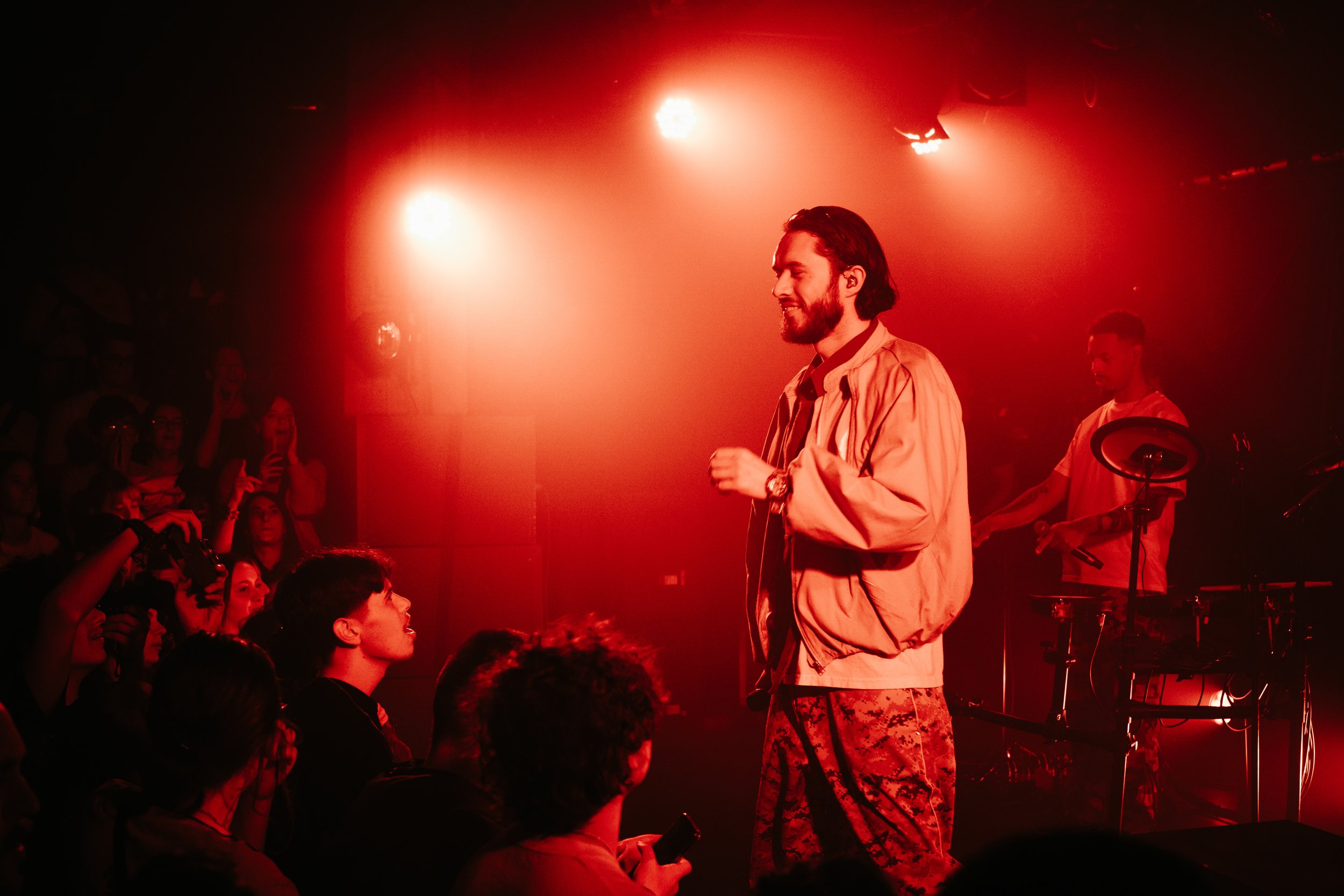 Un chanteur ou rappeur performe sur scène dans une lumière rouge, un public regarde devant avec enthousiasme, un musicien joue en arrière-plan.