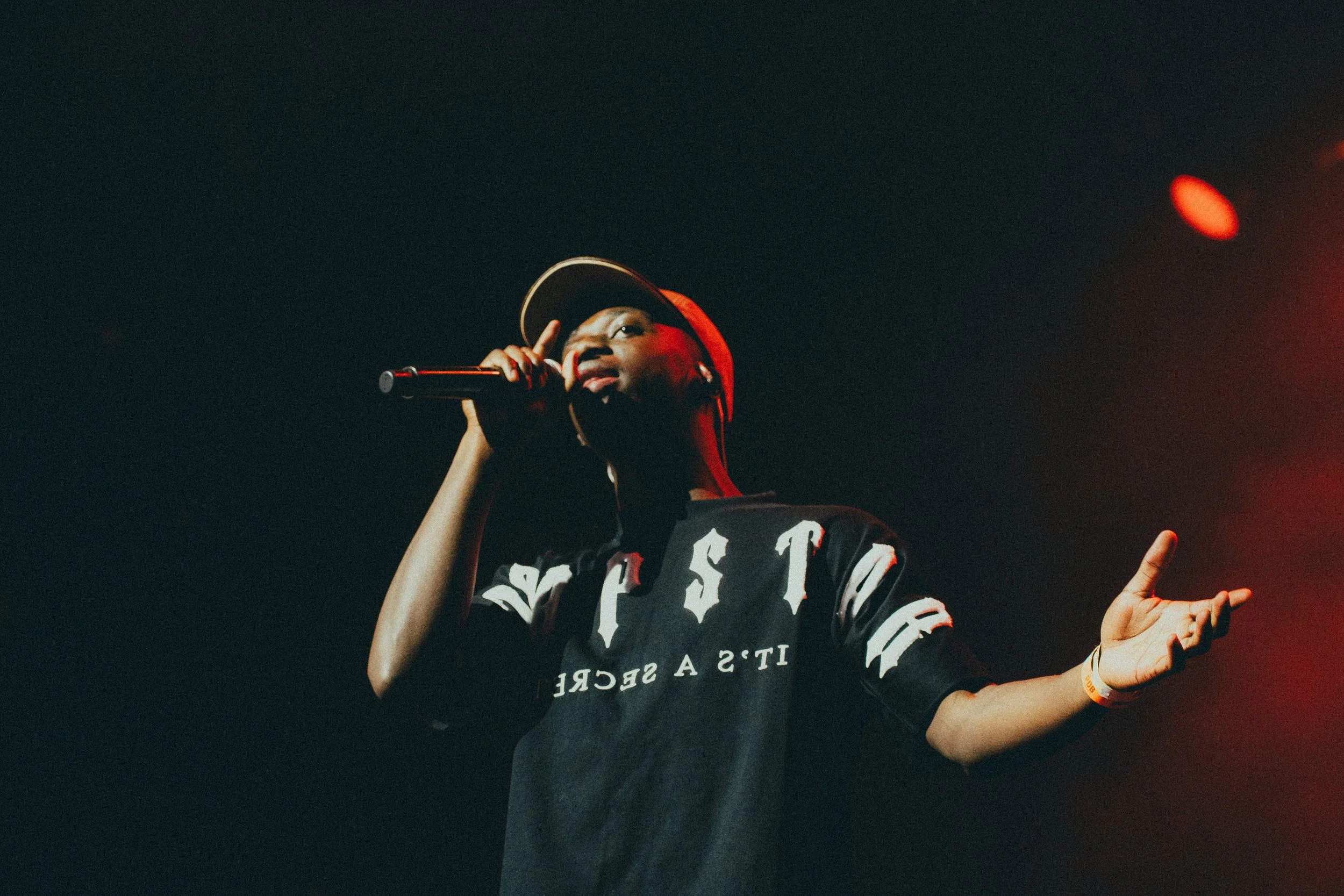 Jeune femme qui chante en micro sur scène, portant un chapeau baseball et un T-shirt noir avec des lettres blanches.