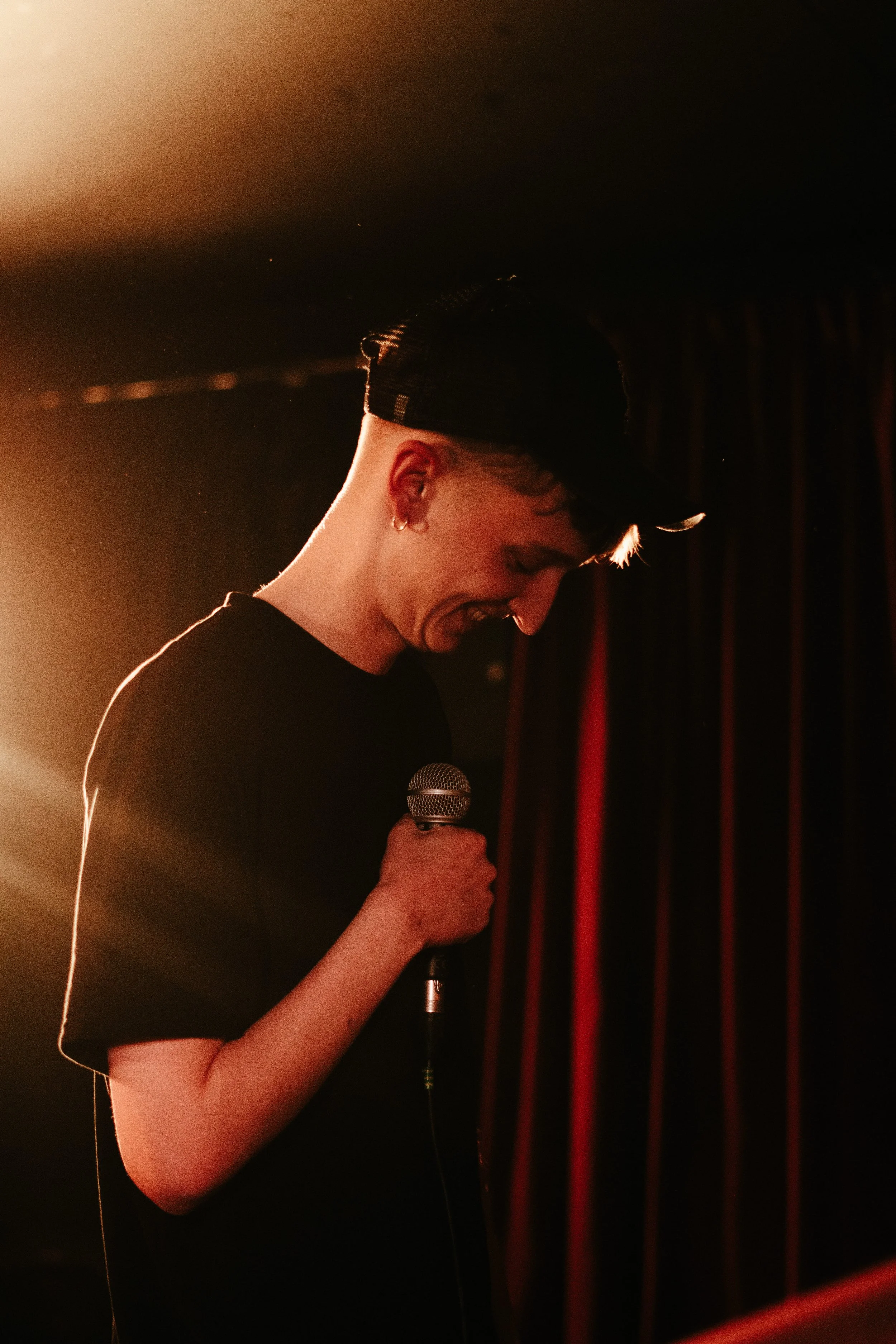 Jeune homme souriant tenant un microphone sur scène avec des rideaux en arrière-plan, éclairé par une lumière chaude.