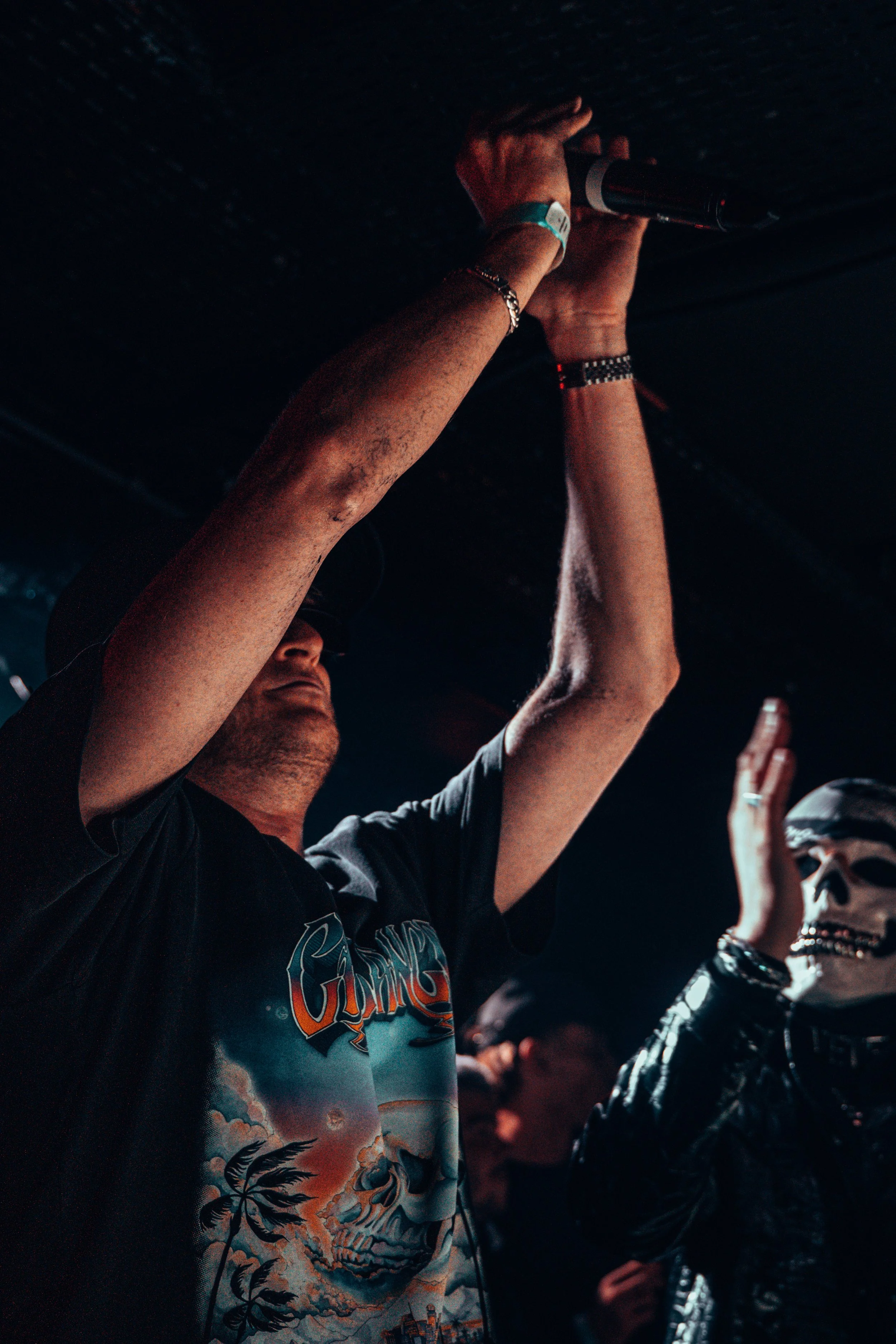 Un homme portant une casquette, un t-shirt avec un dessin de crâne et une scène atmosferique, tient un microphone au plafond lors d'une performance, avec un autre personne portant un masque de squelette en arrière-plan.