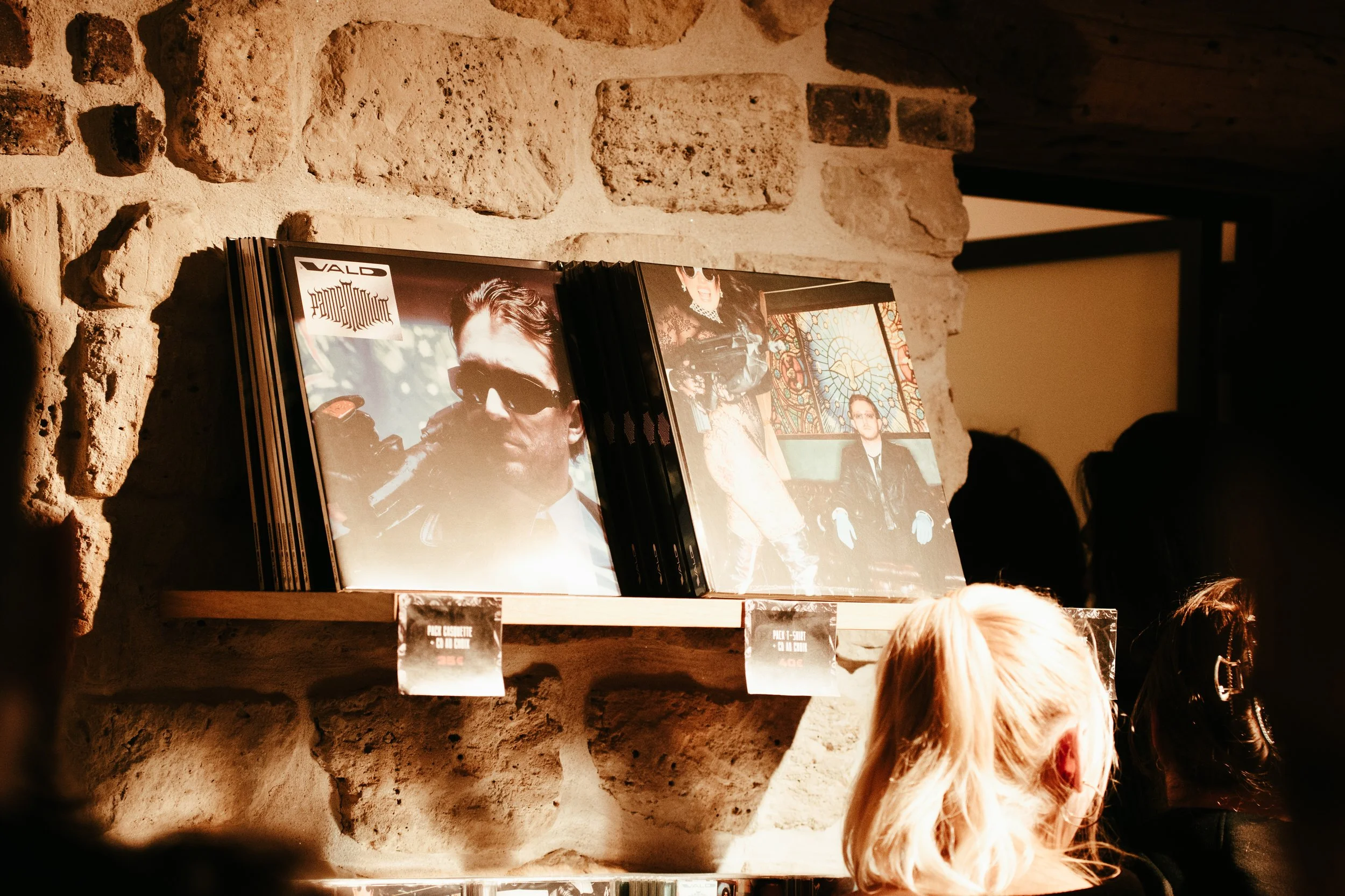 Vinyles exposés sur une étagère en bois contre un mur en pierre, avec plusieurs personnes en arrière-plan dans une ambiance chaleureuse.