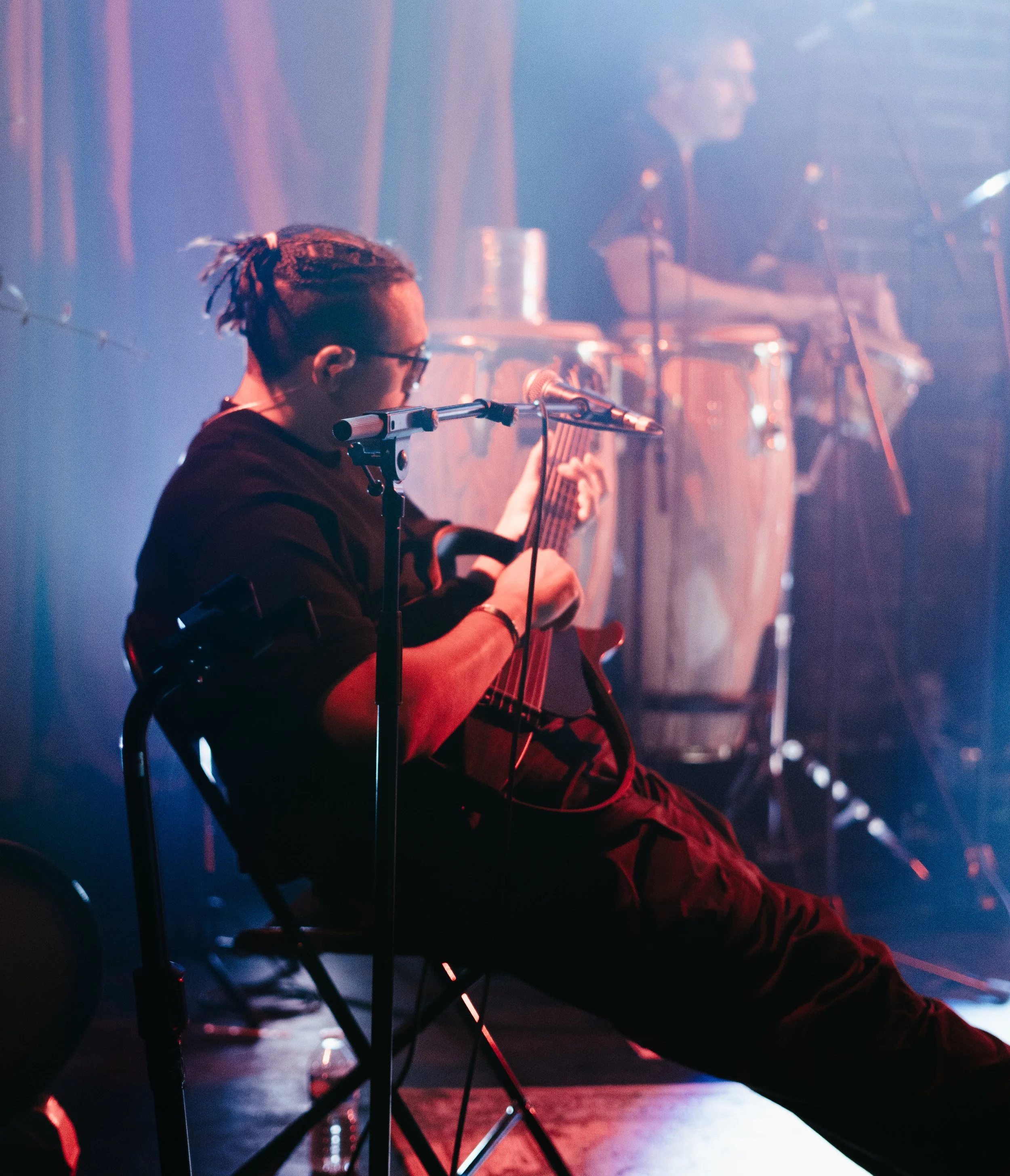 Un musicien joue de la guitare devant une scène avec des instruments de percussion, dans une ambiance tamisée avec des lumières colorées.