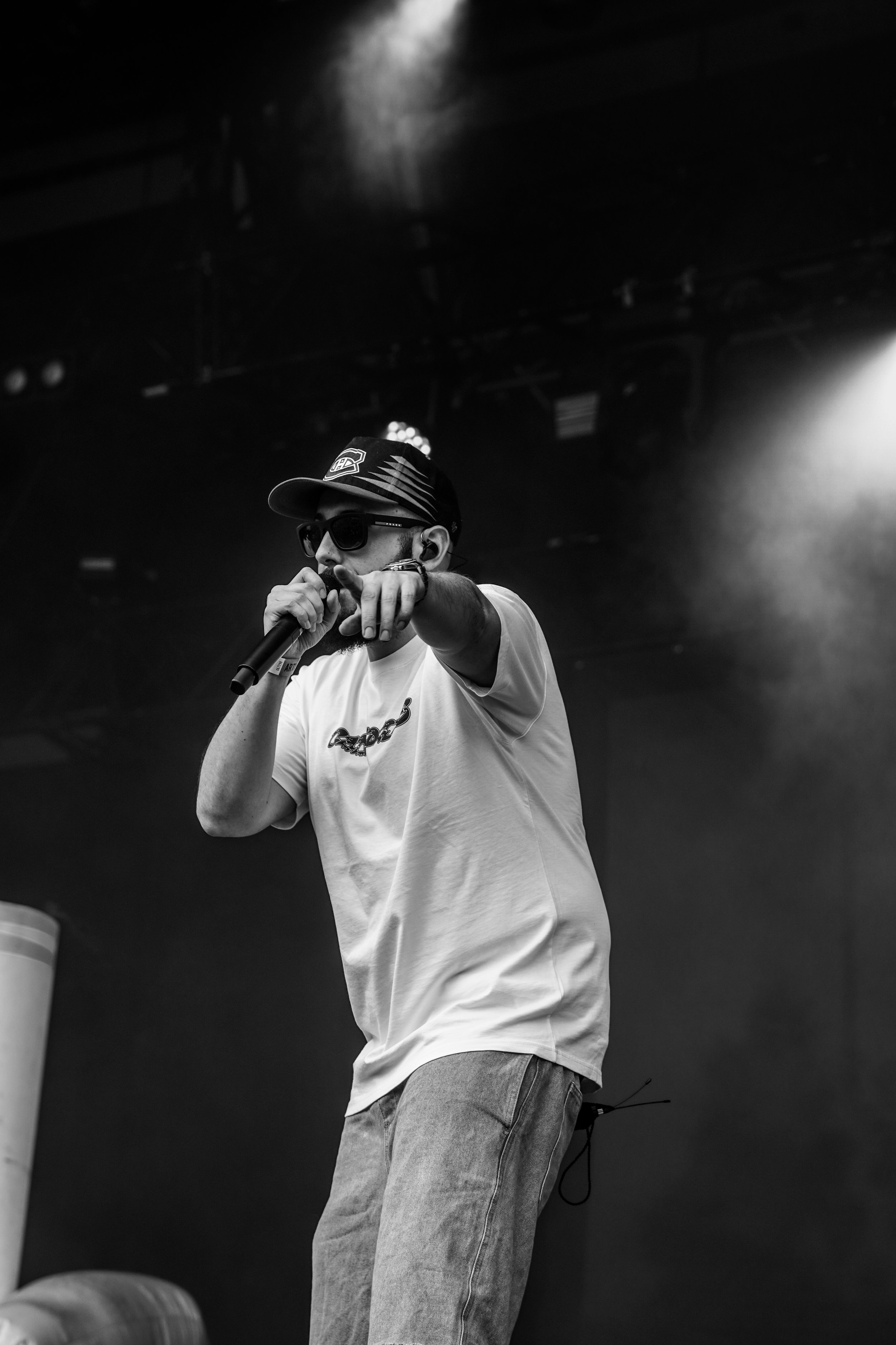 Un homme portant un t-shirt blanc, un chapeau à visière et des lunettes de soleil, tient un micro et semble faire un spectacle sur scène, en noir et blanc.