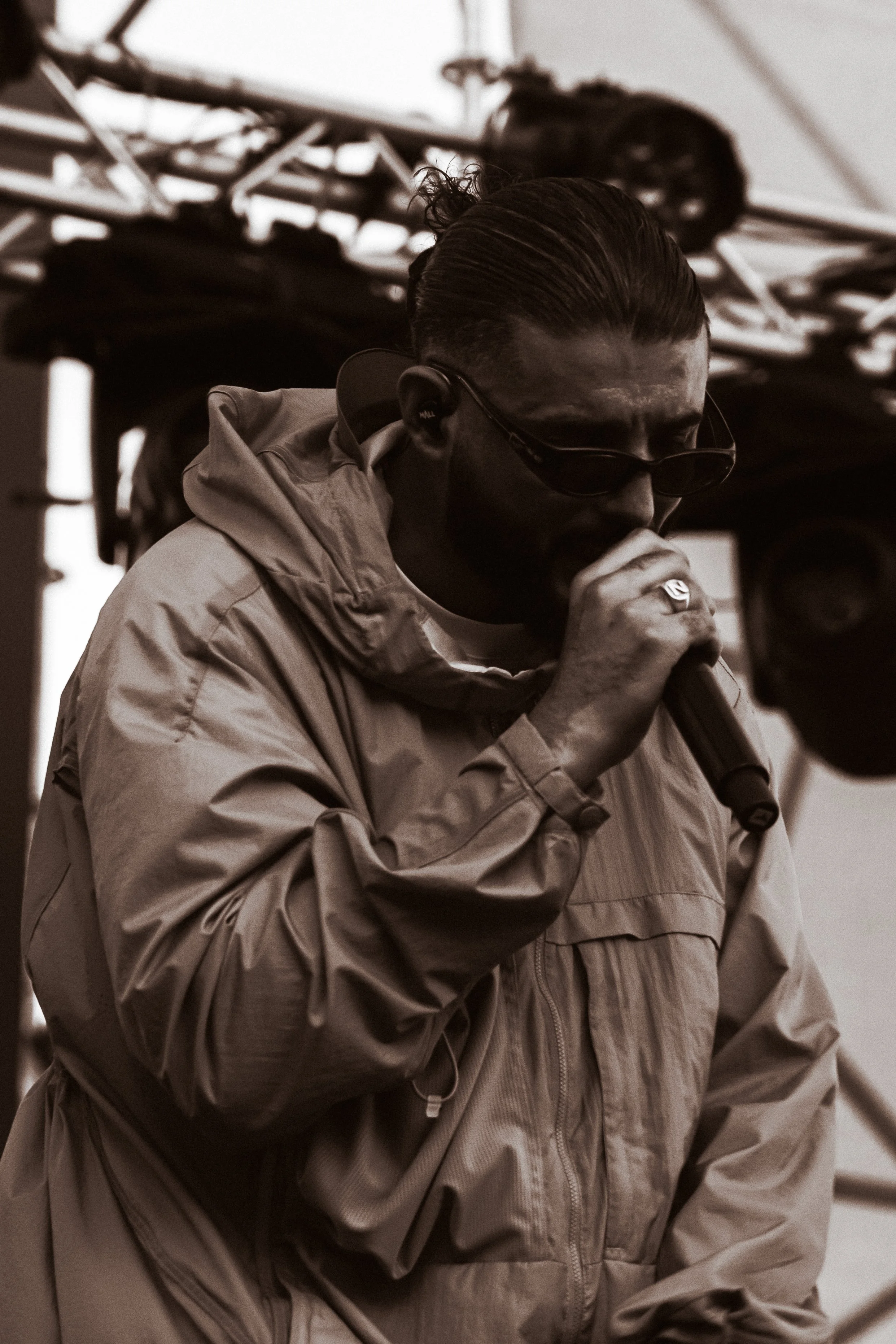 Un homme portant une veste beige, des lunettes de soleil, un casque d'écoute et parlant dans un microphone lors d'un événement en plein air.