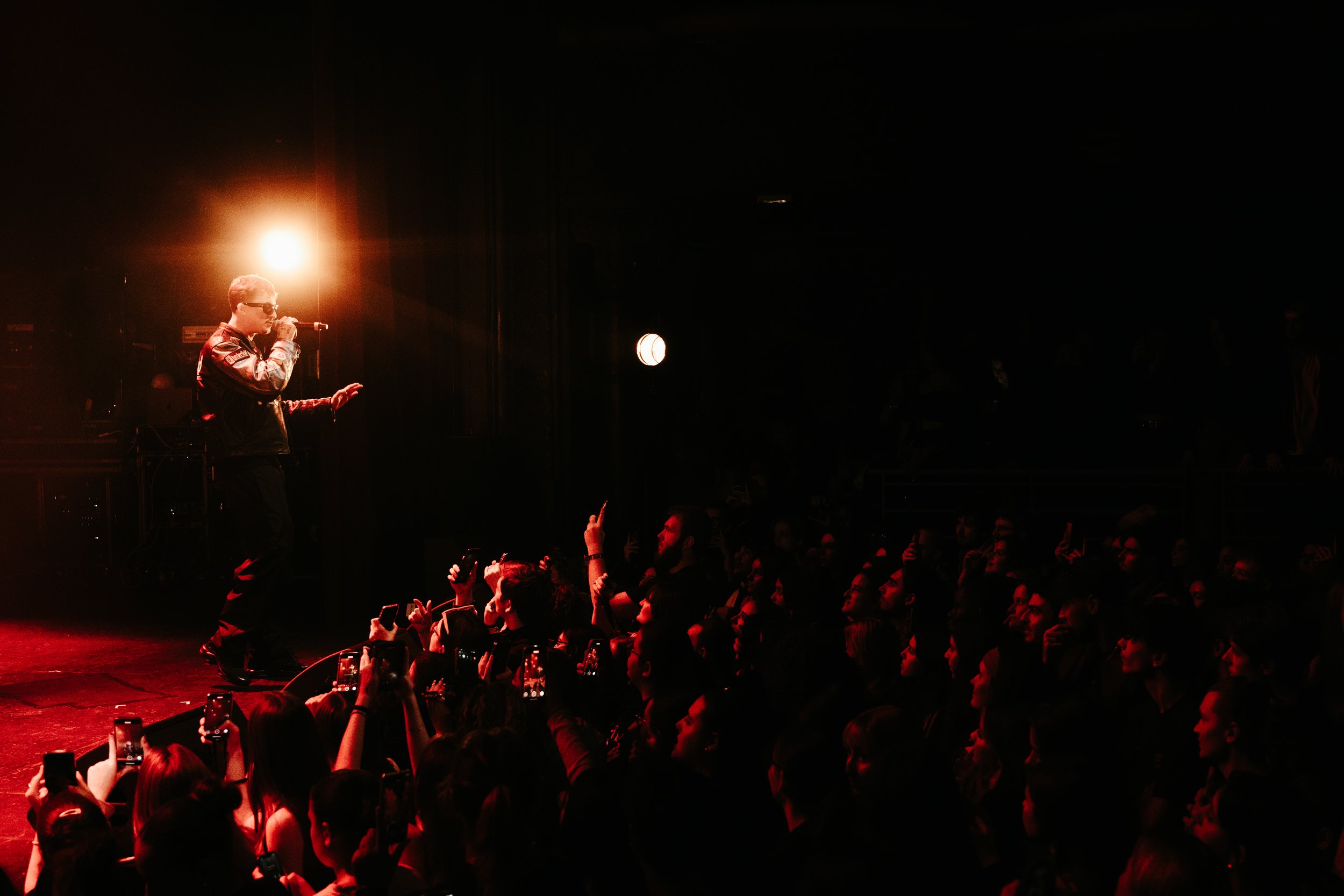 Un chanteur portant des lunettes de soleil et un blouson en cuir, chantant lors d'un concert, avec un public nombreux tenant leurs téléphones pour prendre des photos et vidéos, dans une ambiance sombre mais avec un éclairage puissant sur l'artiste.