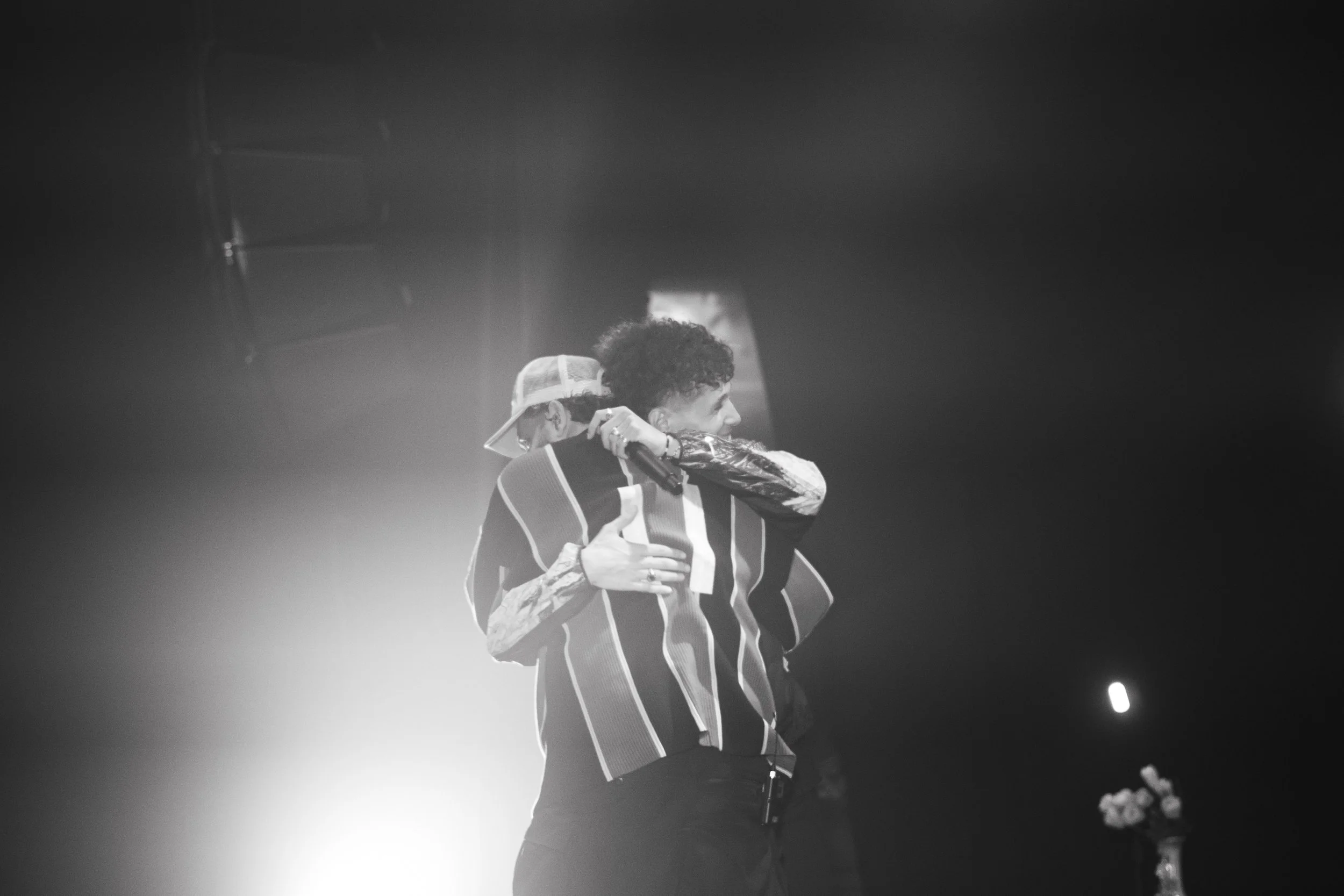 Deux personnes s'embrassent sur scène, l'une porte une casquette blanche et l'autre a des cheveux bouclés, sous un éclairage sombre.