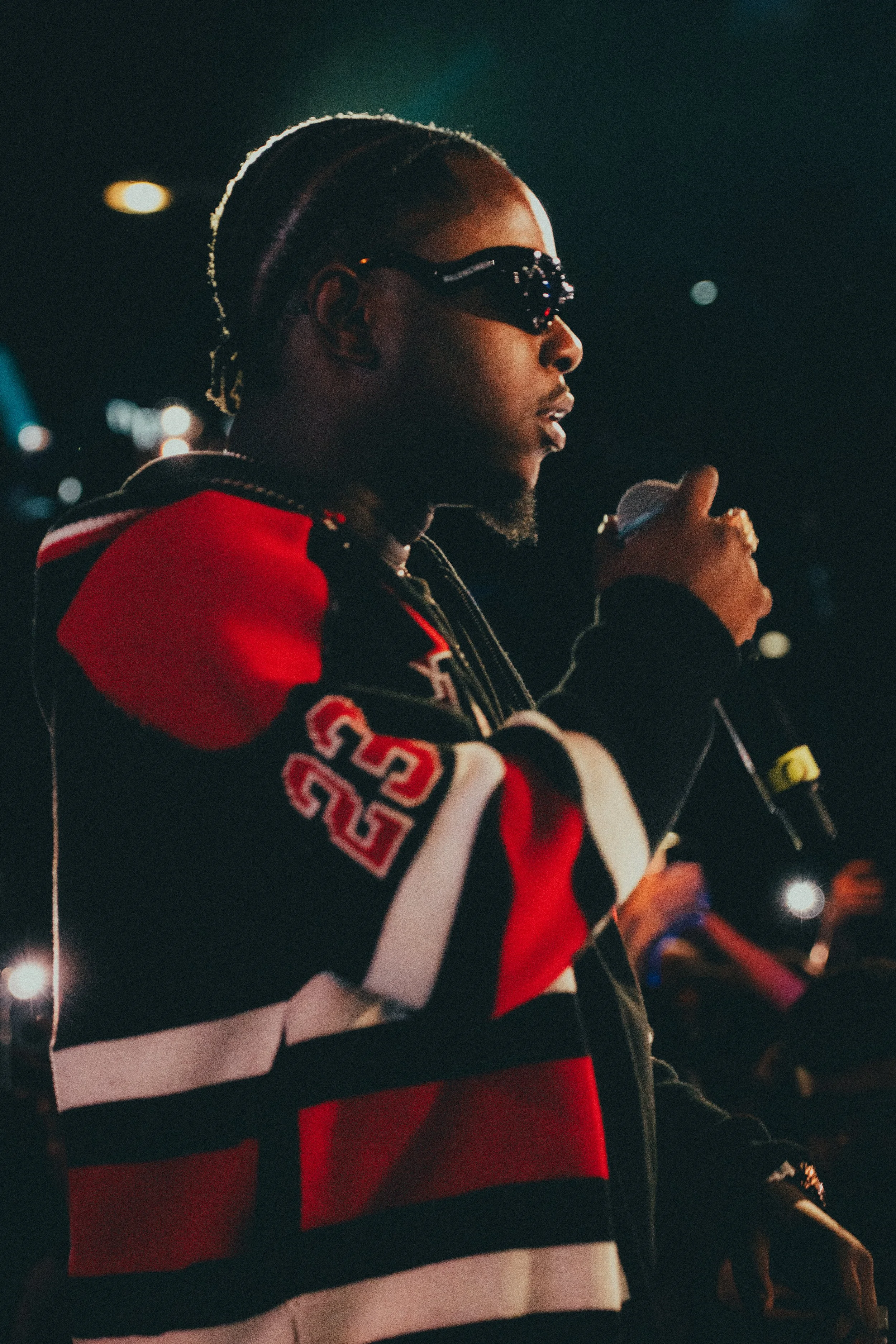 Un homme avec des cheveux tressés portant des lunettes noires et un sweat à rayures rouges, noires et blanches, tenant un microphone lors d'une performance ou d'une présentation dans un environnement sombre.