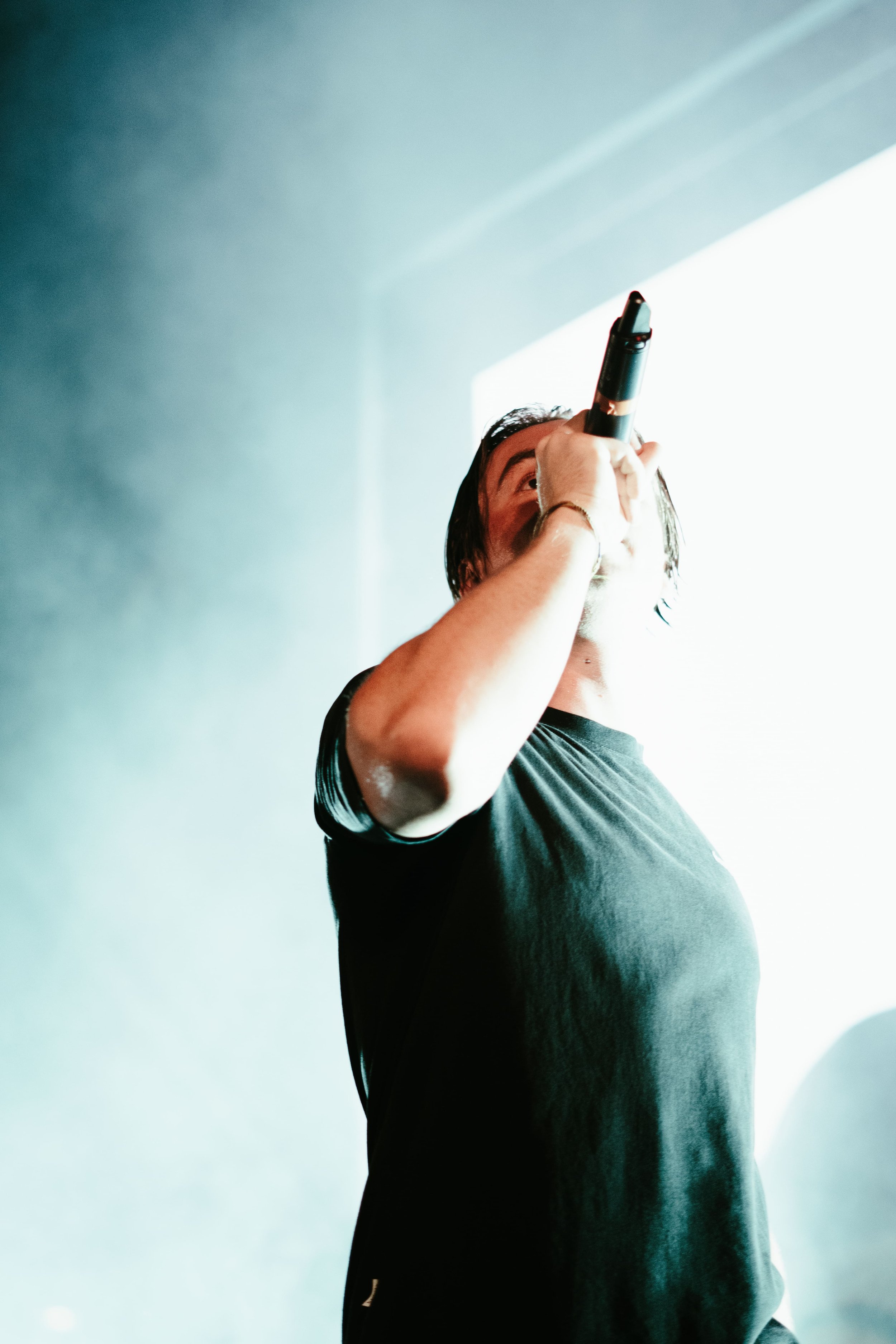 Un homme chantant ou parlant dans un microphone, vu de profil, sur scène ou dans une ambiance sombre avec une lumière vive en arrière-plan.