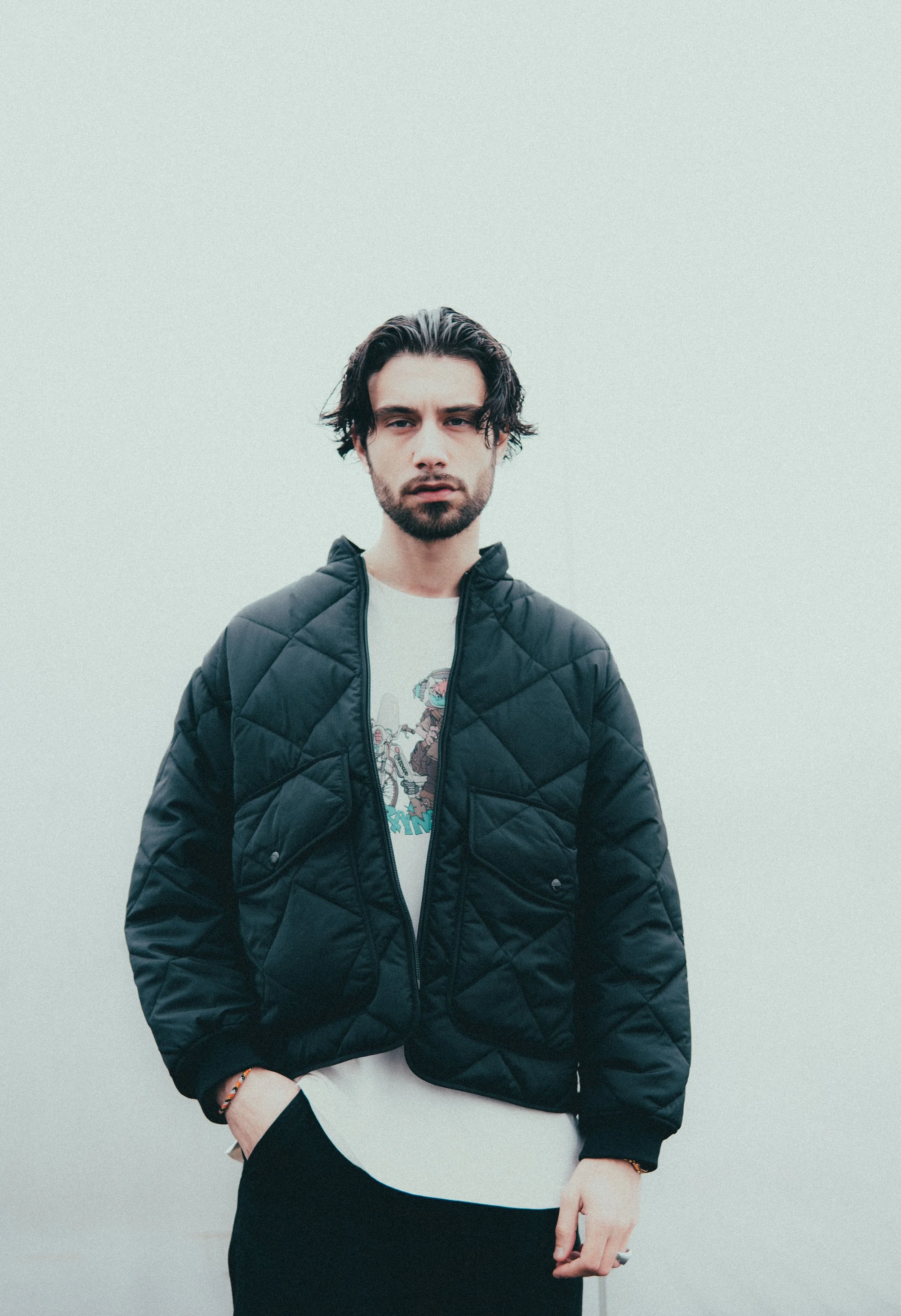 Un jeune homme avec des cheveux longs et une barbe, portant une veste noire, une chemise blanche et un t-shirt graphique, pose devant un fond gris clair.