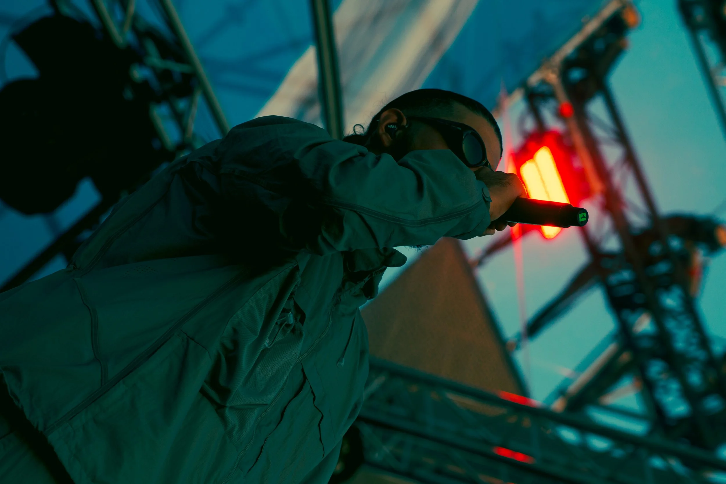 Un homme portant des lunettes et une veste regarde une lumière rouge avec un microphone dans la main dans un environnement urbain nocturne.