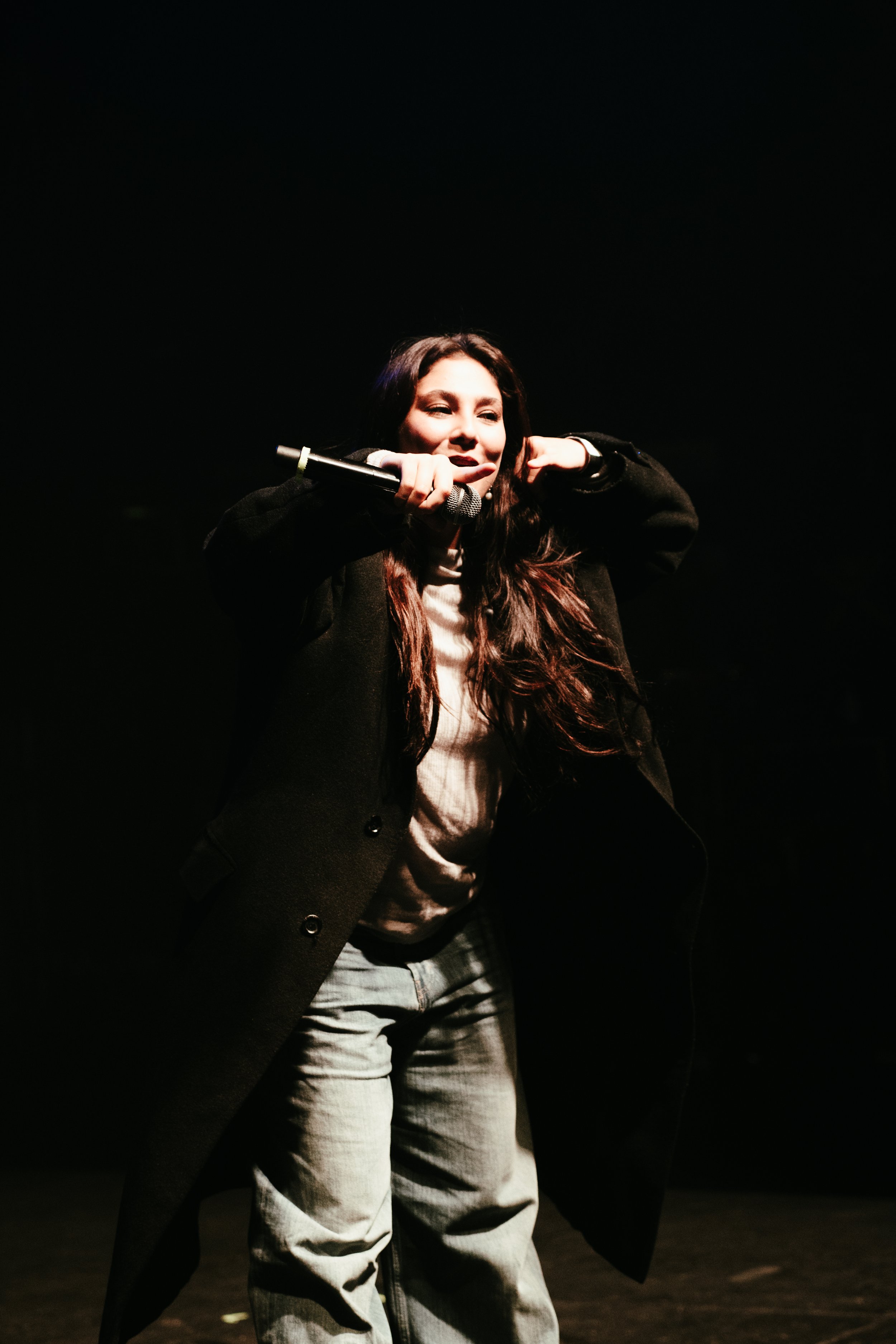 Une jeune femme avec de longs cheveux bruns, portant un manteau noir, tient un microphone et semble en train de performer sur scène dans un éclairage tamisé.