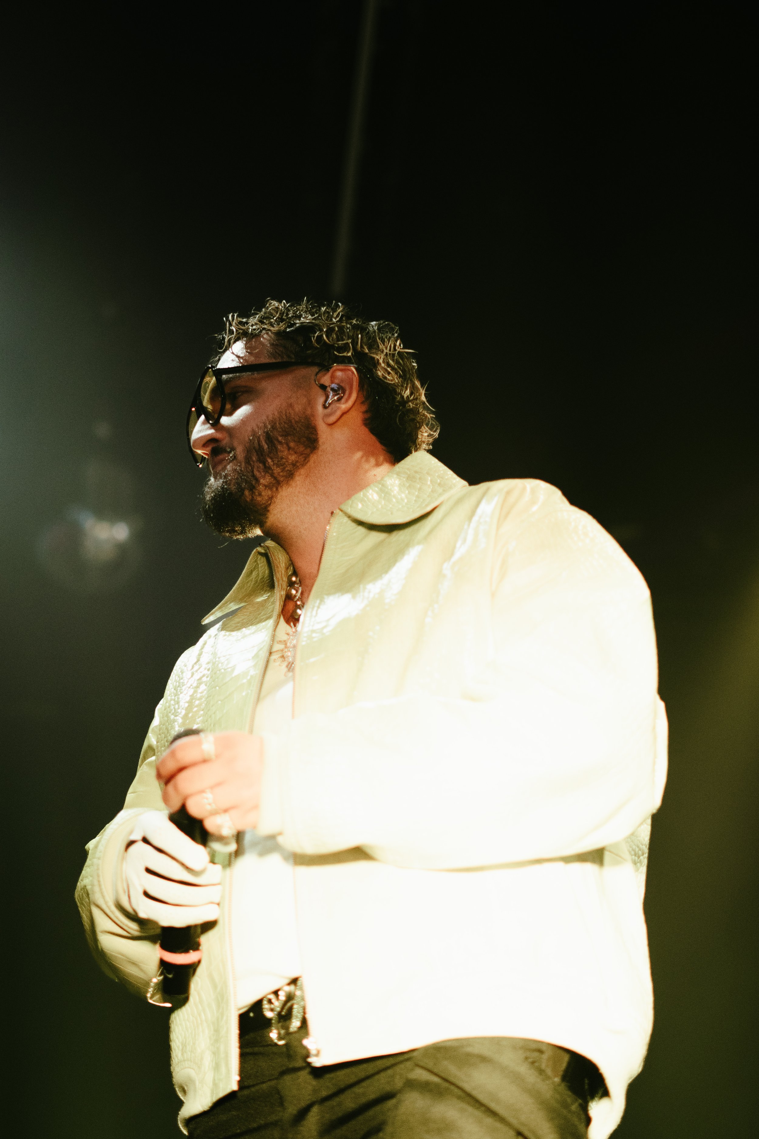 Un homme chantant sur scène, portant des lunettes de soleil, un casque d'écoute, une veste crème et tenant un microphone.