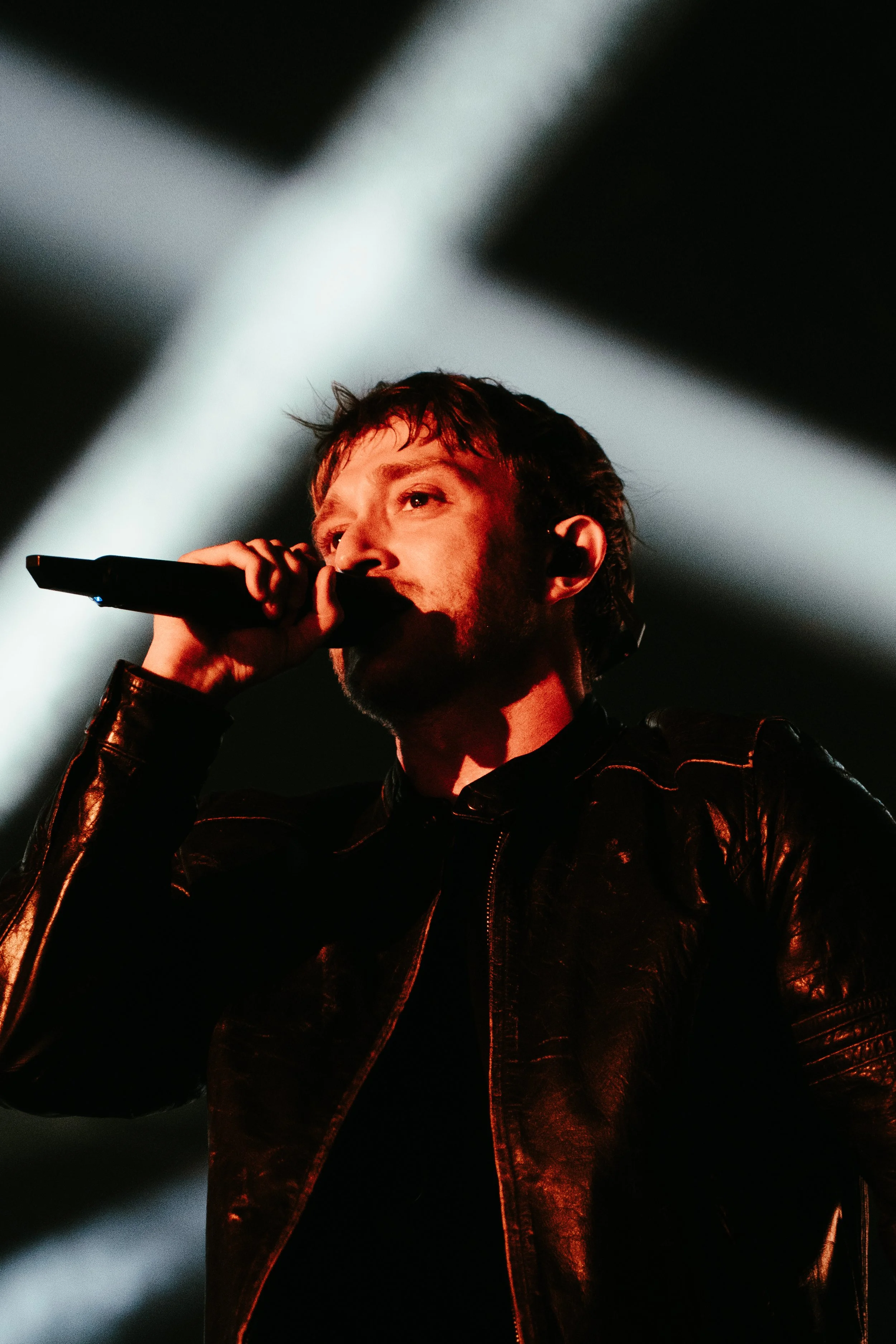 Homme chantant avec un micro sur scène, portant une veste en cuir noire, éclairé par des projecteurs lumineux.