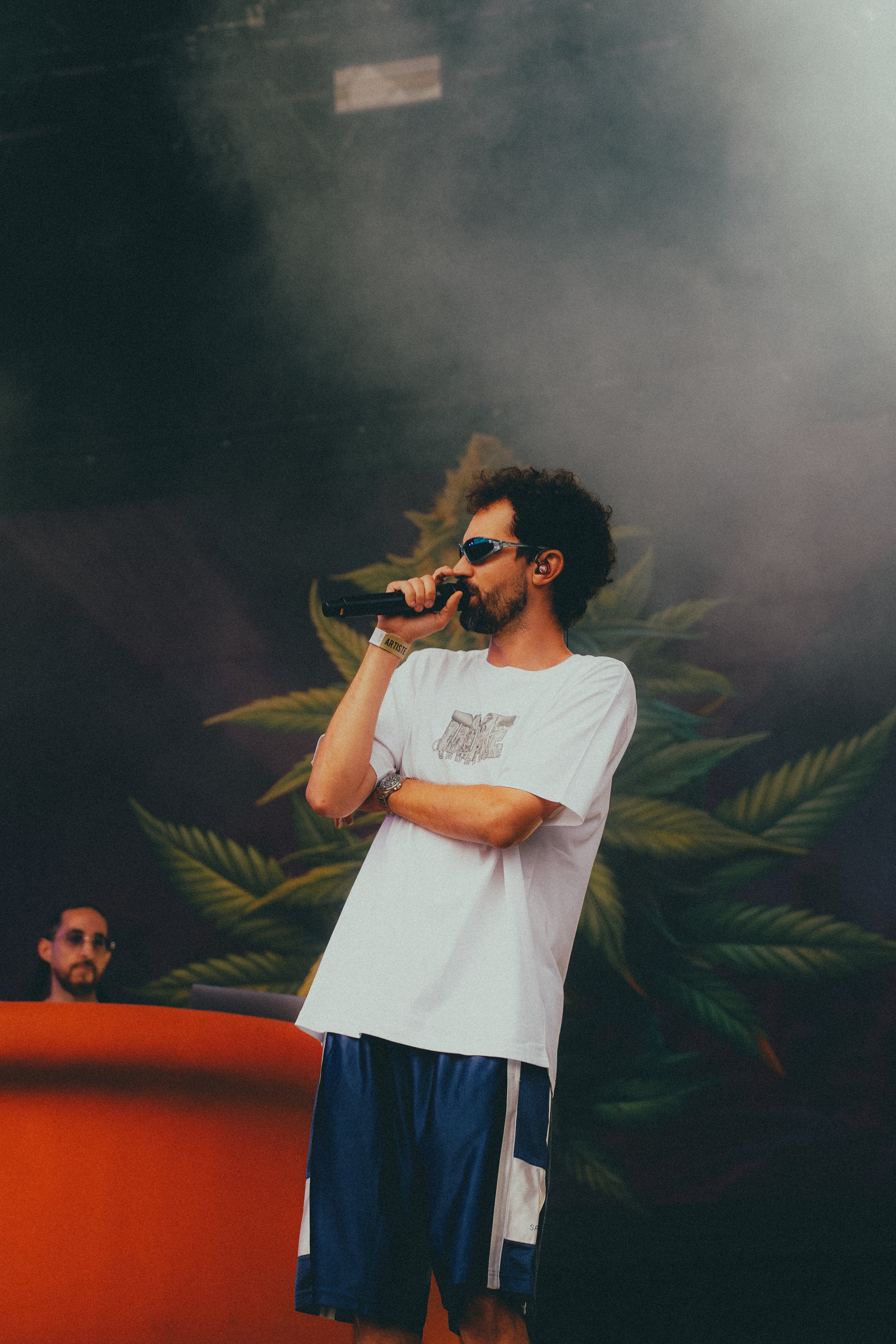 Un homme avec des cheveux frisés et une barbe, portant des lunettes de soleil, un t-shirt blanc et un short de sport, tient un micro lors d'une performance sur scène. En arrière-plan, un autre homme avec des lunettes de soleil et une barbe est devant