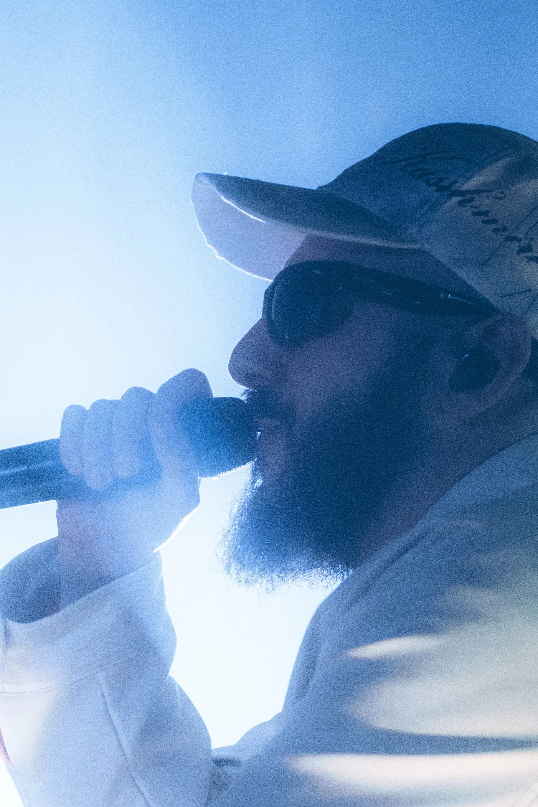 Homme portant une casquette, lunettes de soleil, chantant dans un microphone, avec un fond lumineux.