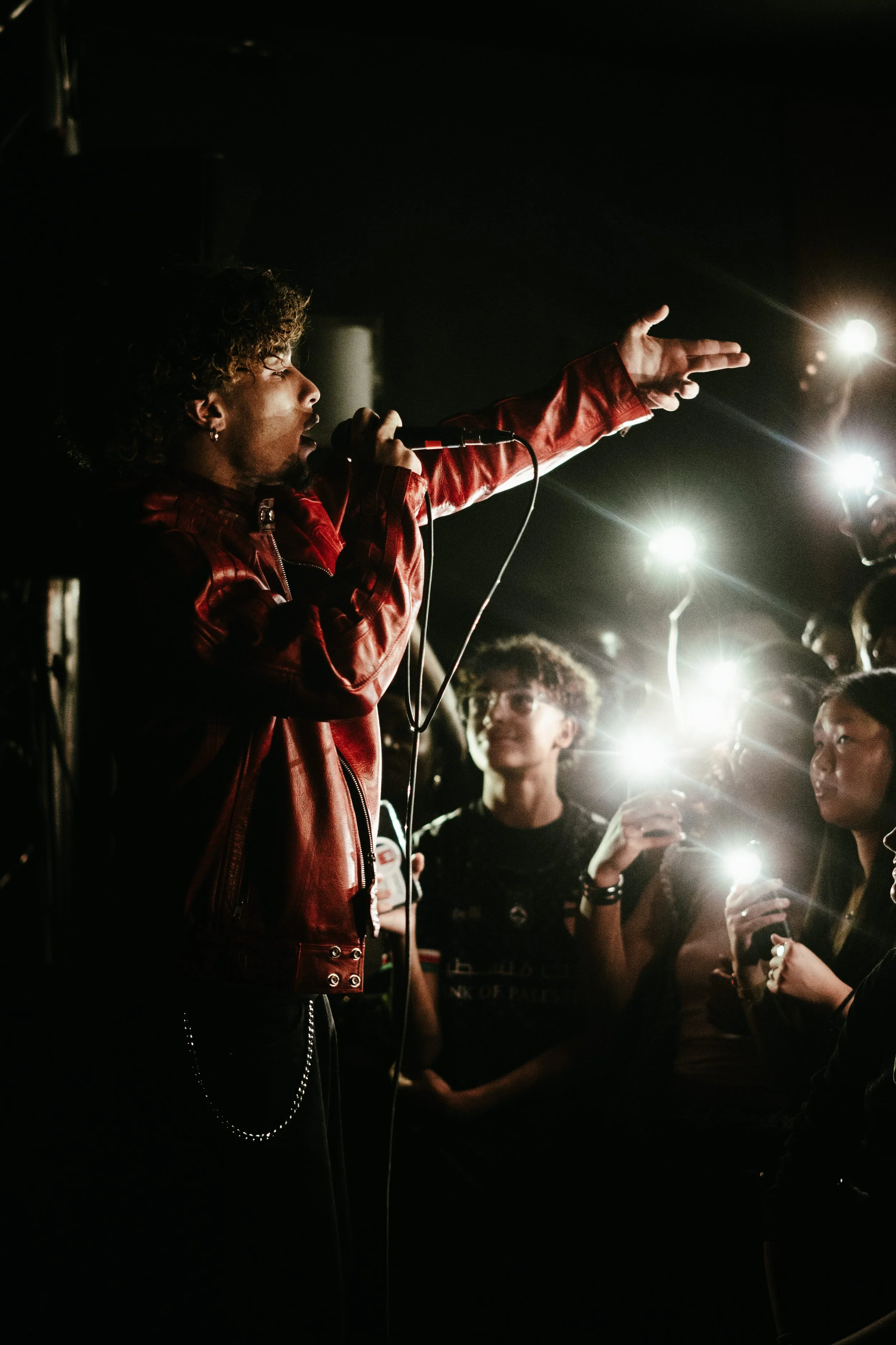 Un chanteur portant une veste en cuir rouge sur scène, s'adressant à un public avec des lumières brillantes. Le public tient des téléphones avec torches allumées.
