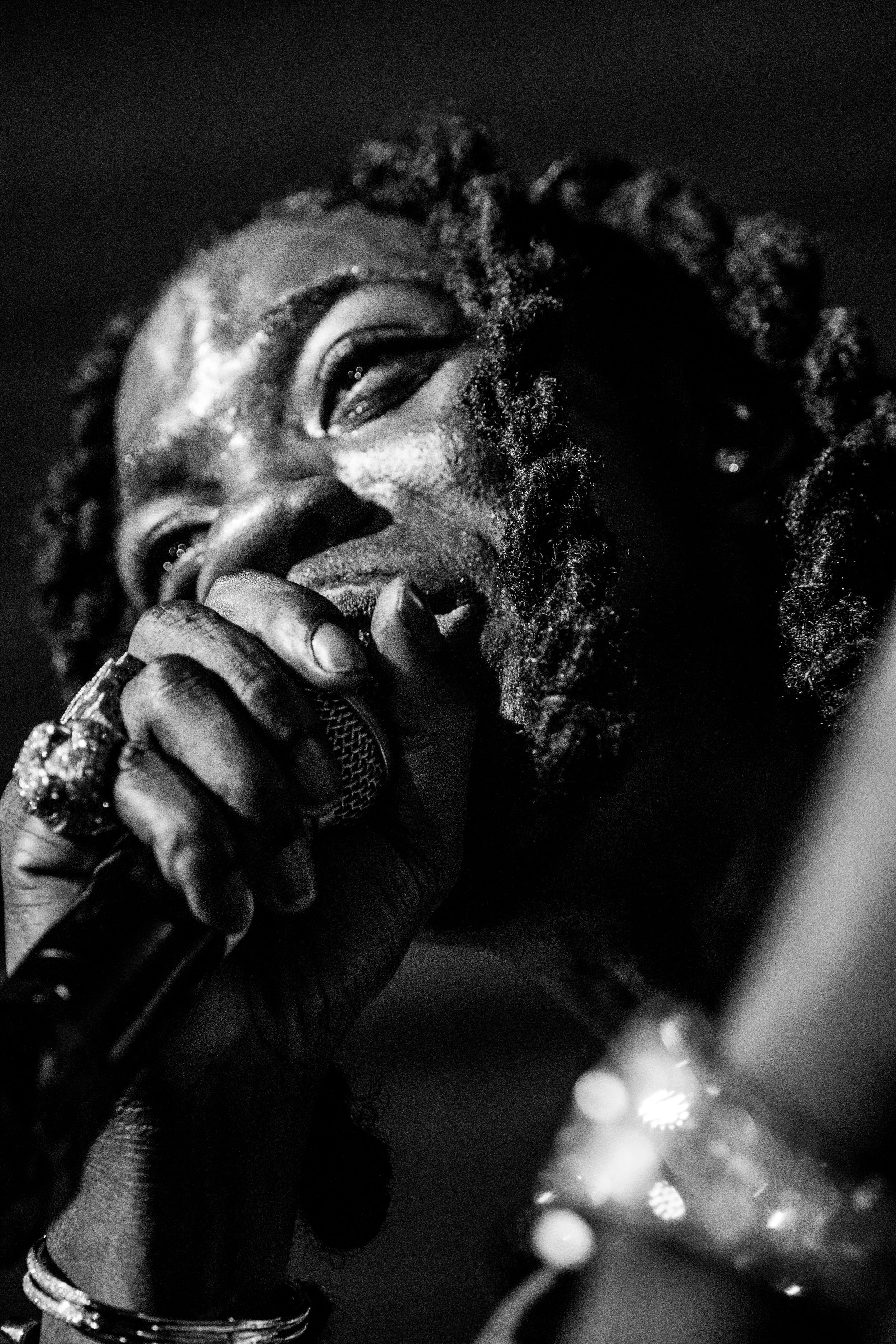 Un homme noir avec des cheveux crépus, tenant un microphone et semblant chanter ou parler, en noir et blanc.