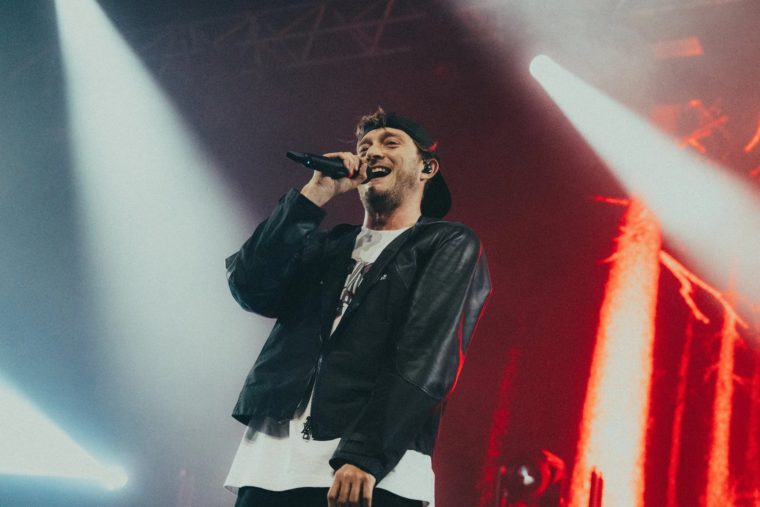 Un homme chante avec un micro lors d'un concert, portant une veste en cuir noir et un chapeau noir à l'envers, avec un éclairage de scène en arrière-plan.