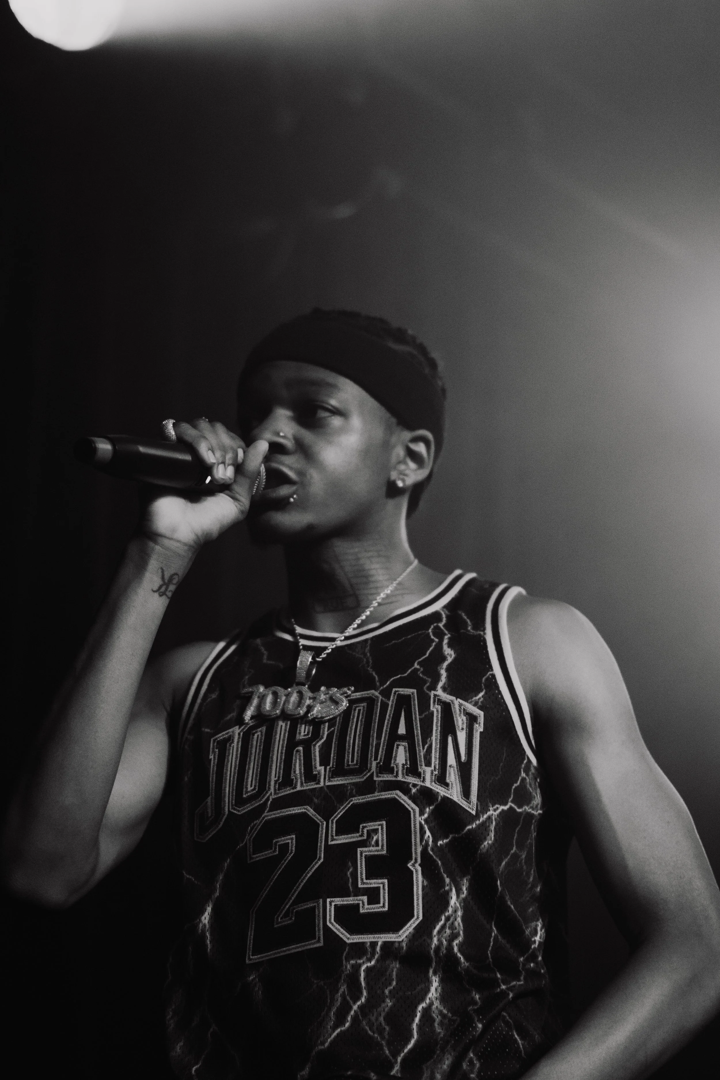 Jeune femme noire en t-shirt Jordan 23, portant un bandeau noir, tenant un micro en chantant ou parlant sur scène en noir et blanc.