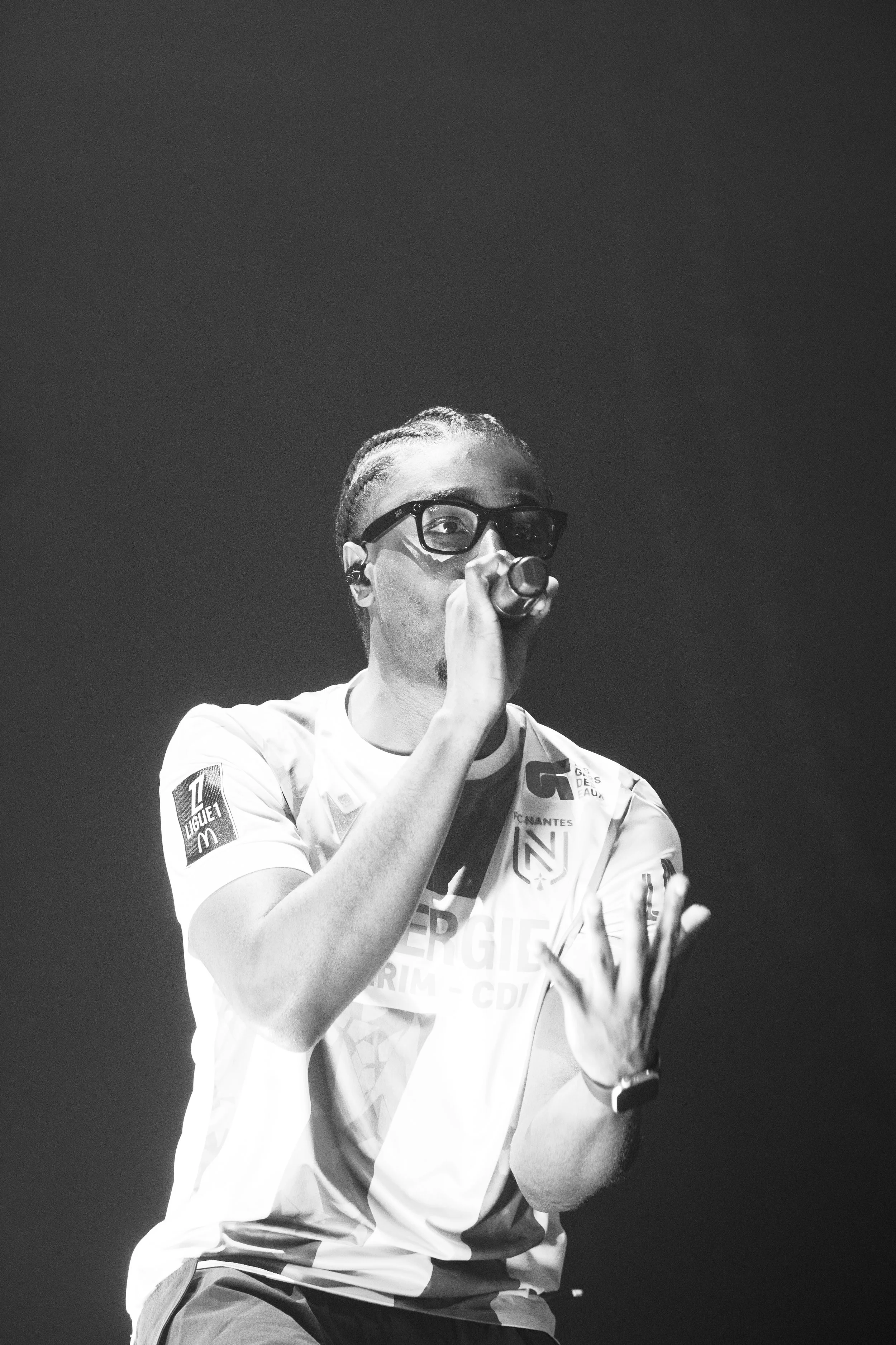 Une personne portant des lunettes et un maillot de football chante en tenant un microphone dans une main et levant la main de l'autre.