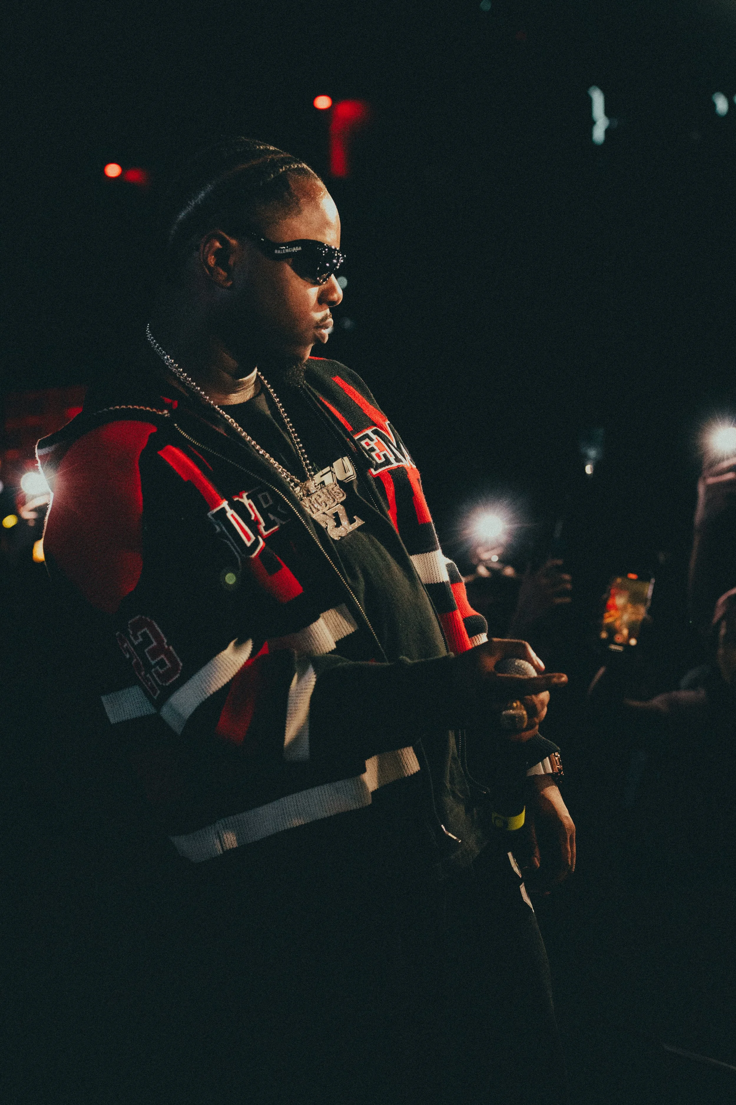 Une personne portant des lunettes de soleil et une veste à rayures rouges, noires et blanches, tient un téléphone portable dans un environnement sombre avec des lumières brillantes en arrière-plan.